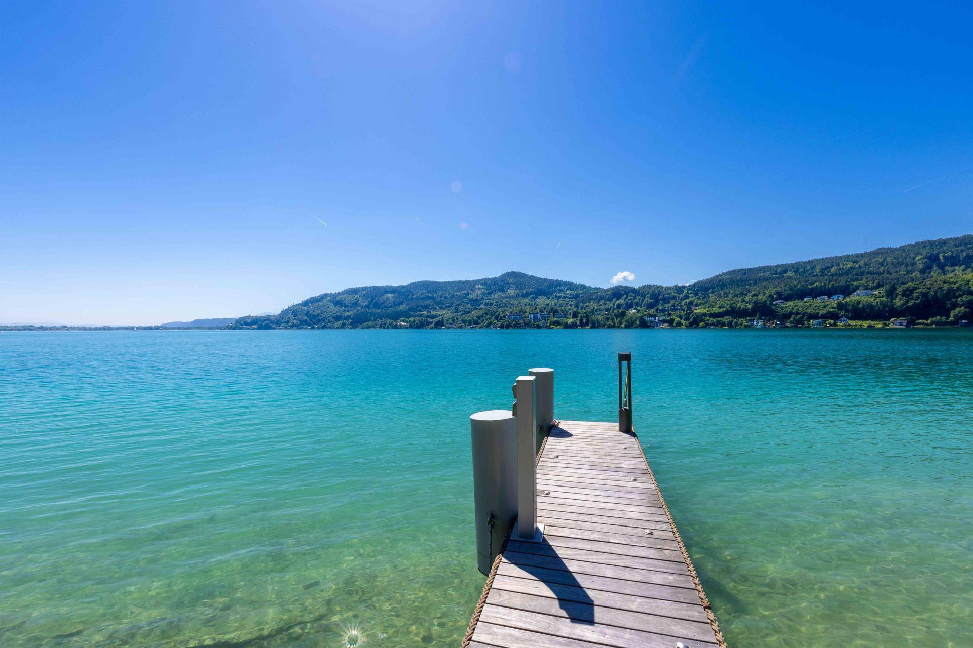 Blick vom Ende des Holzstegs auf das weite, türkisfarbene Seewasser und die bewaldeten Berge.