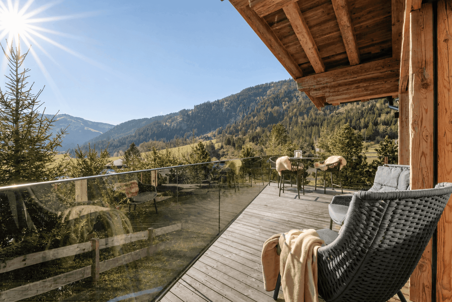 Geräumiger Holzbalkon mit Glasgeländer, bequemen Sitzgelegenheiten und Panoramablick auf die Alpen.