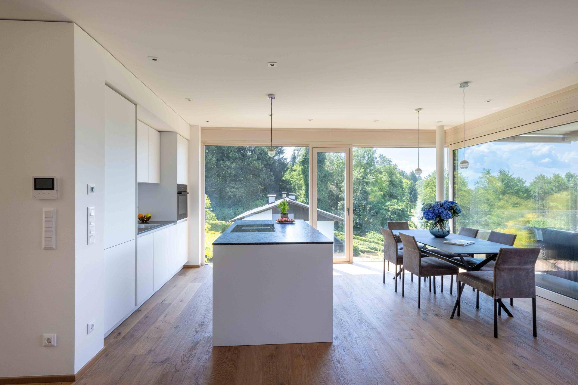Helle, moderne Küche mit weißer Insel und Essbereich, bietet durch große Fenster einen Blick ins Grüne.