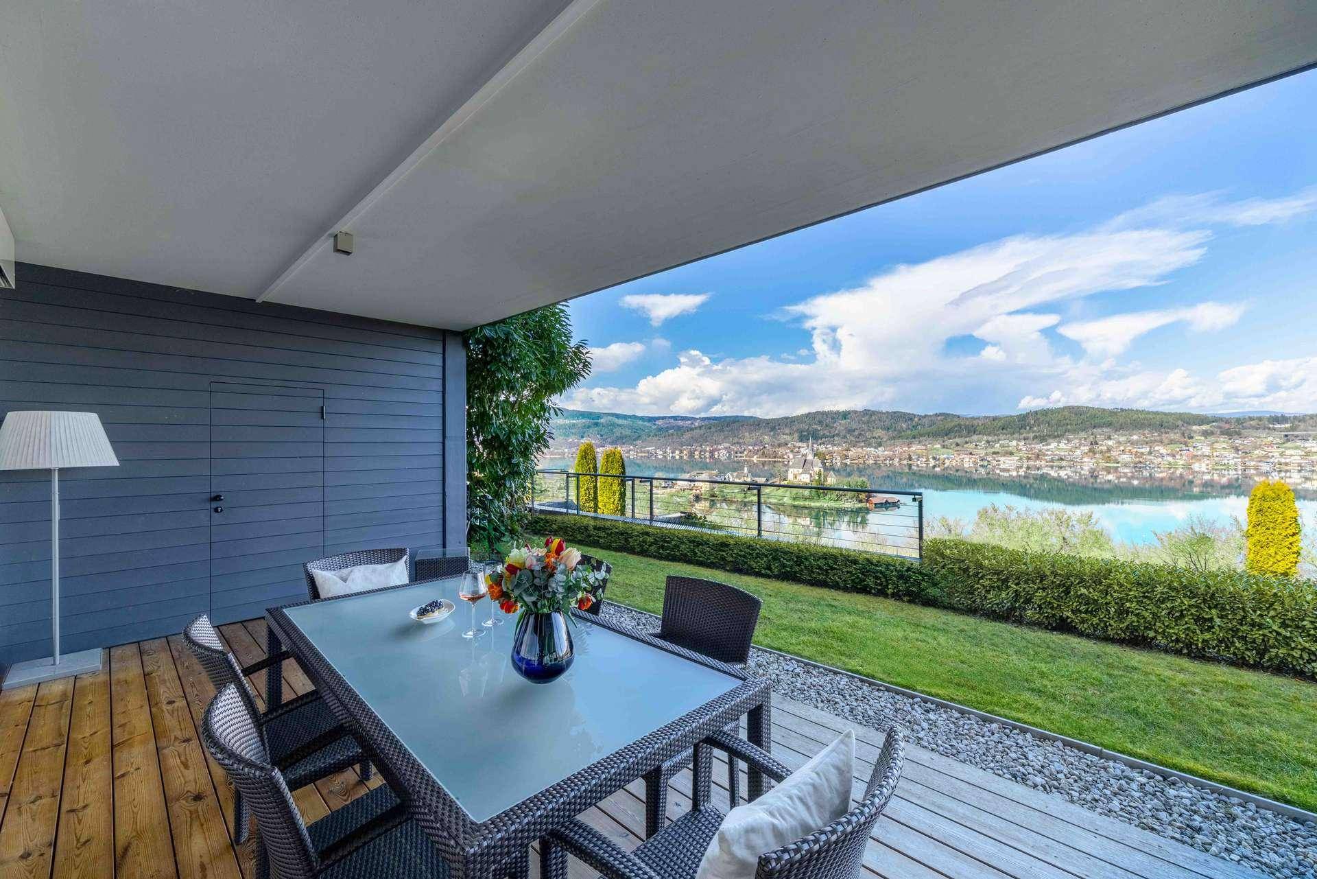 Überdachte Terrasse mit Esstisch und Seeblick, umgeben von Rasen und Hecken.