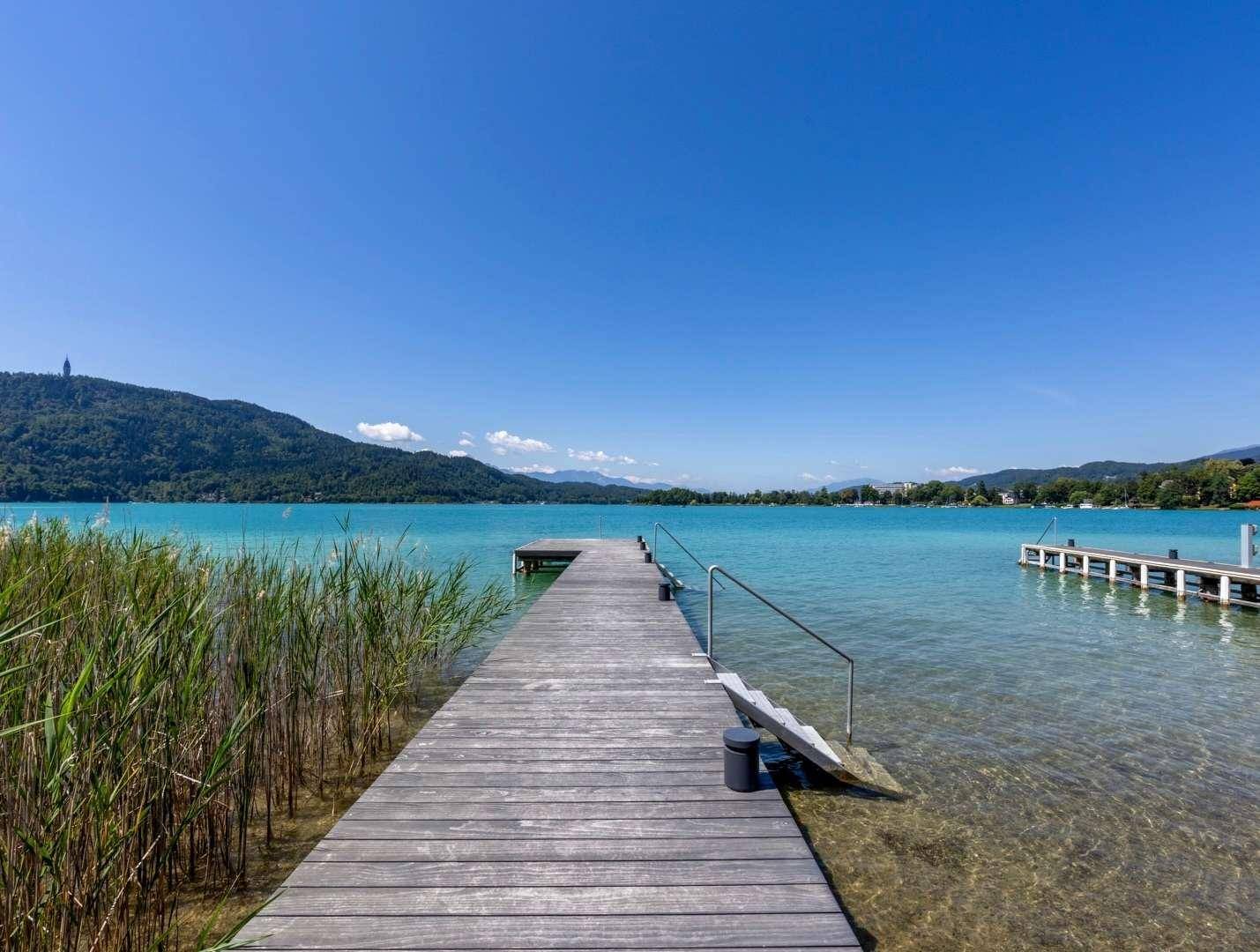 Langer Holzsteg führt ins klare, türkisfarbene Wasser des Sees, umgeben von Schilf.