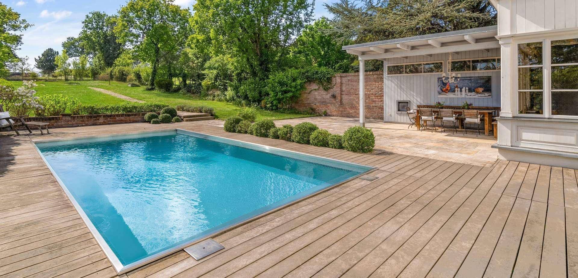 Großzügiger Poolbereich mit Holzdeck, umgeben von Grün und einem überdachten Essplatz im Freien.