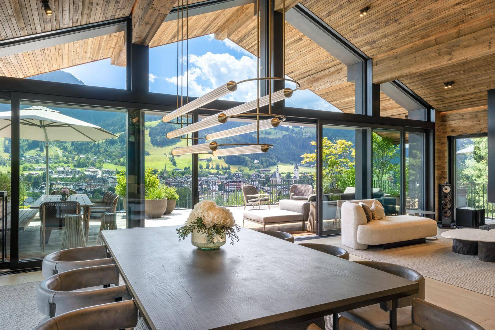Heller Wohn- und Essbereich mit Holzbalkendecke, modernem Kronleuchter und Panoramafenstern mit Bergblick.