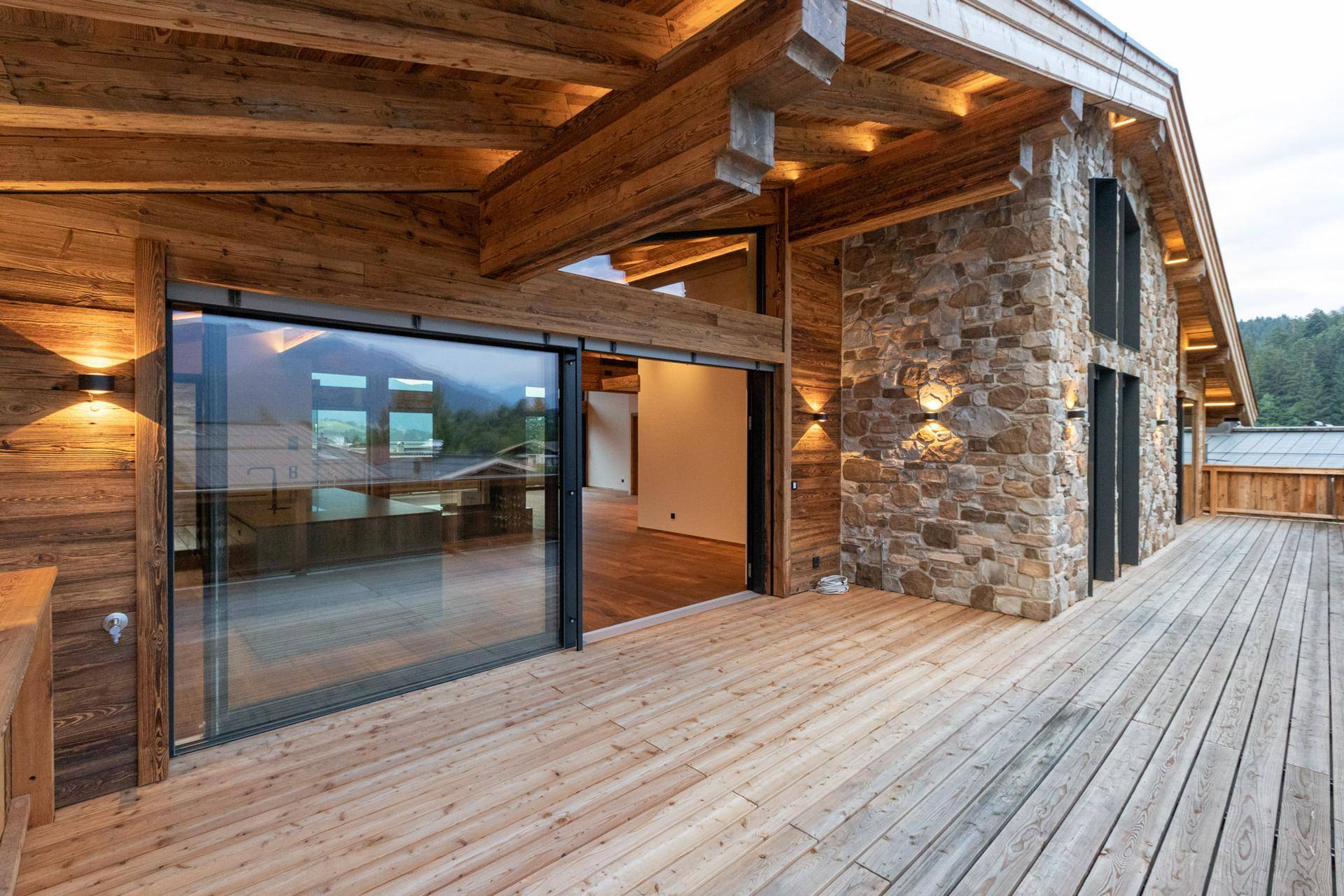 Große Holzterrasse mit Zugang zum Innenbereich und einer Fassade aus Naturstein und Holz.