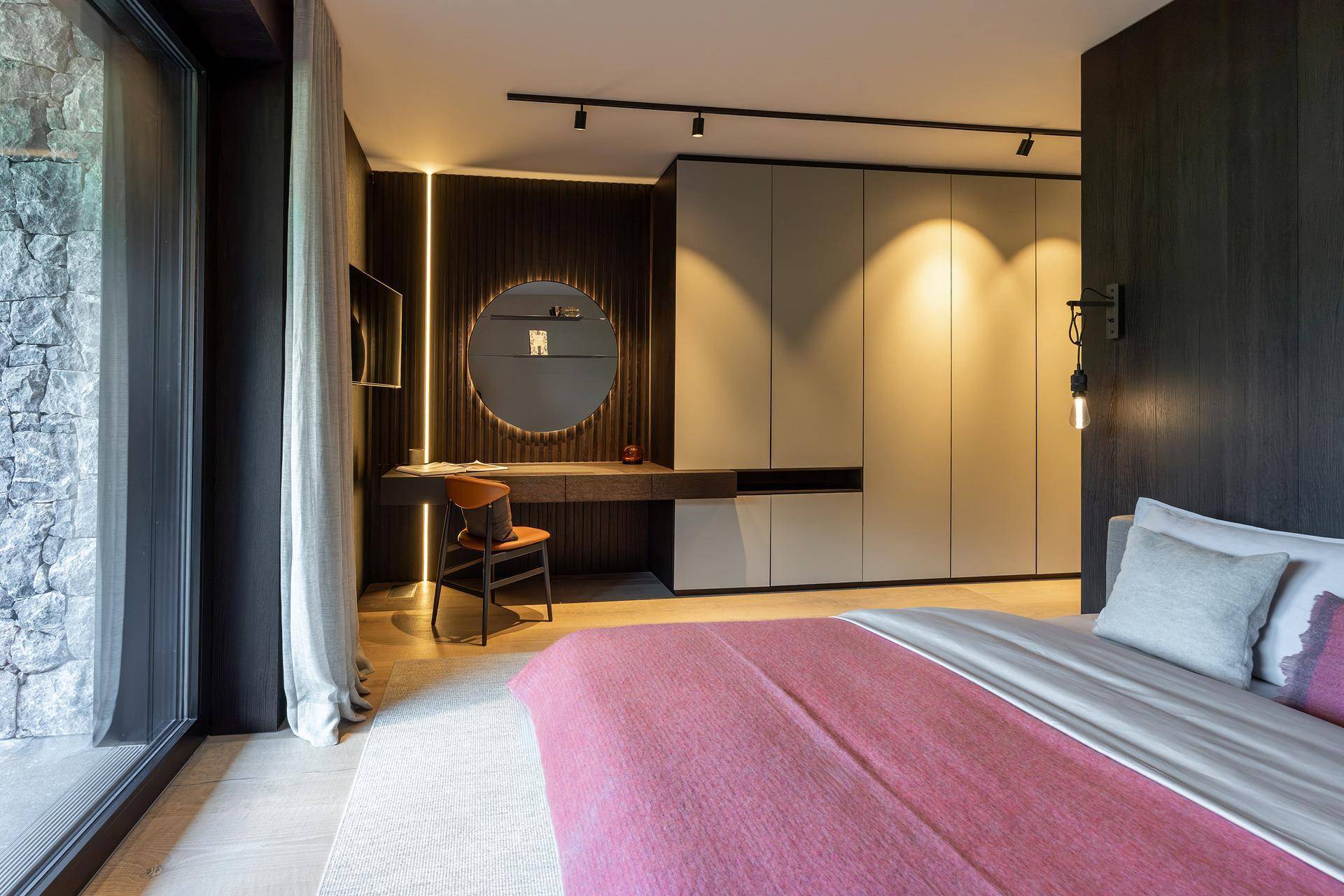Modernes Schlafzimmer mit integriertem Kleiderschrank, Schreibtisch und großem Fenster mit Blick auf eine Steinwand.