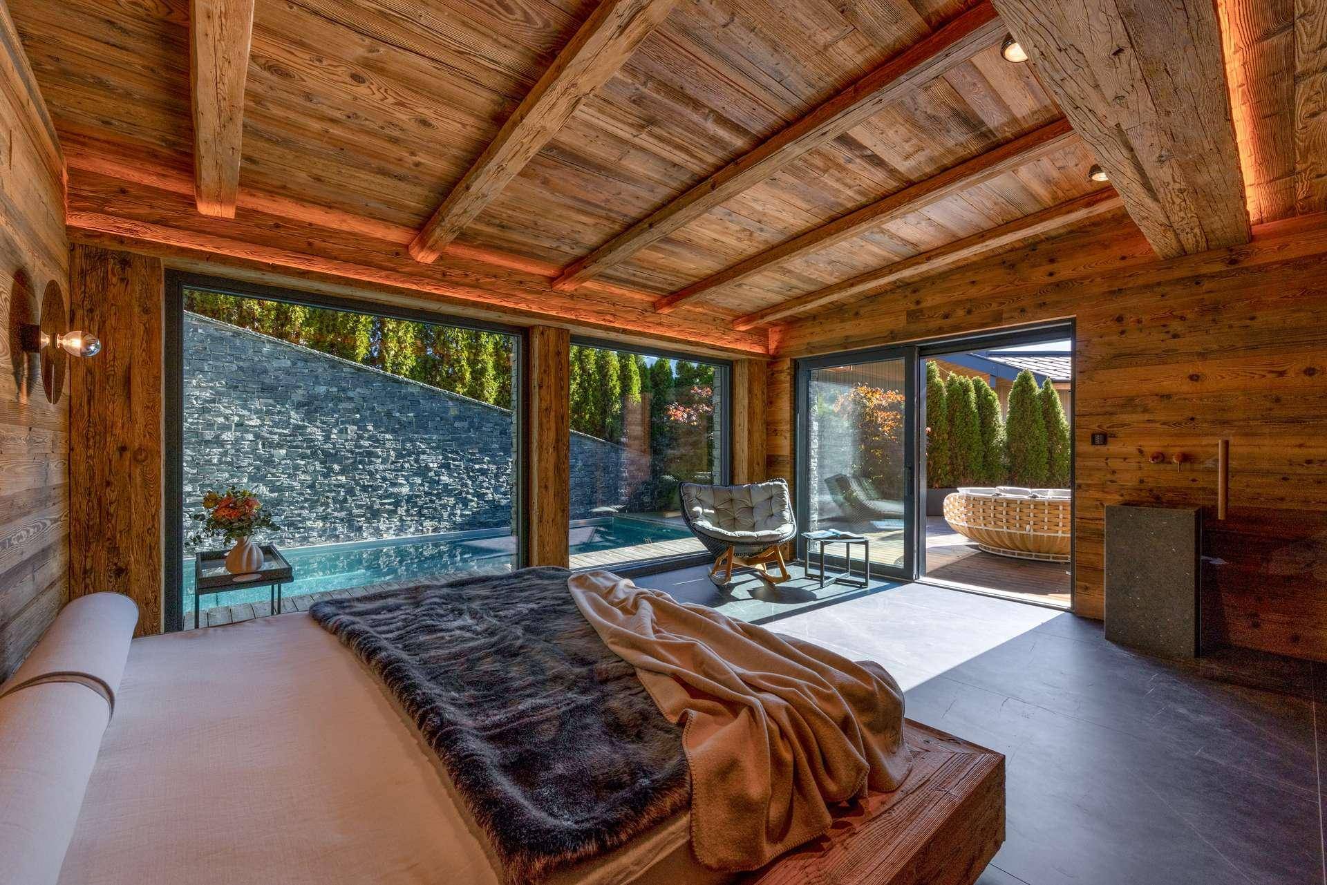 Schlafzimmer mit Holzwänden und großen Fenstern, die den Blick auf einen Außenpool und die Natur freigeben.