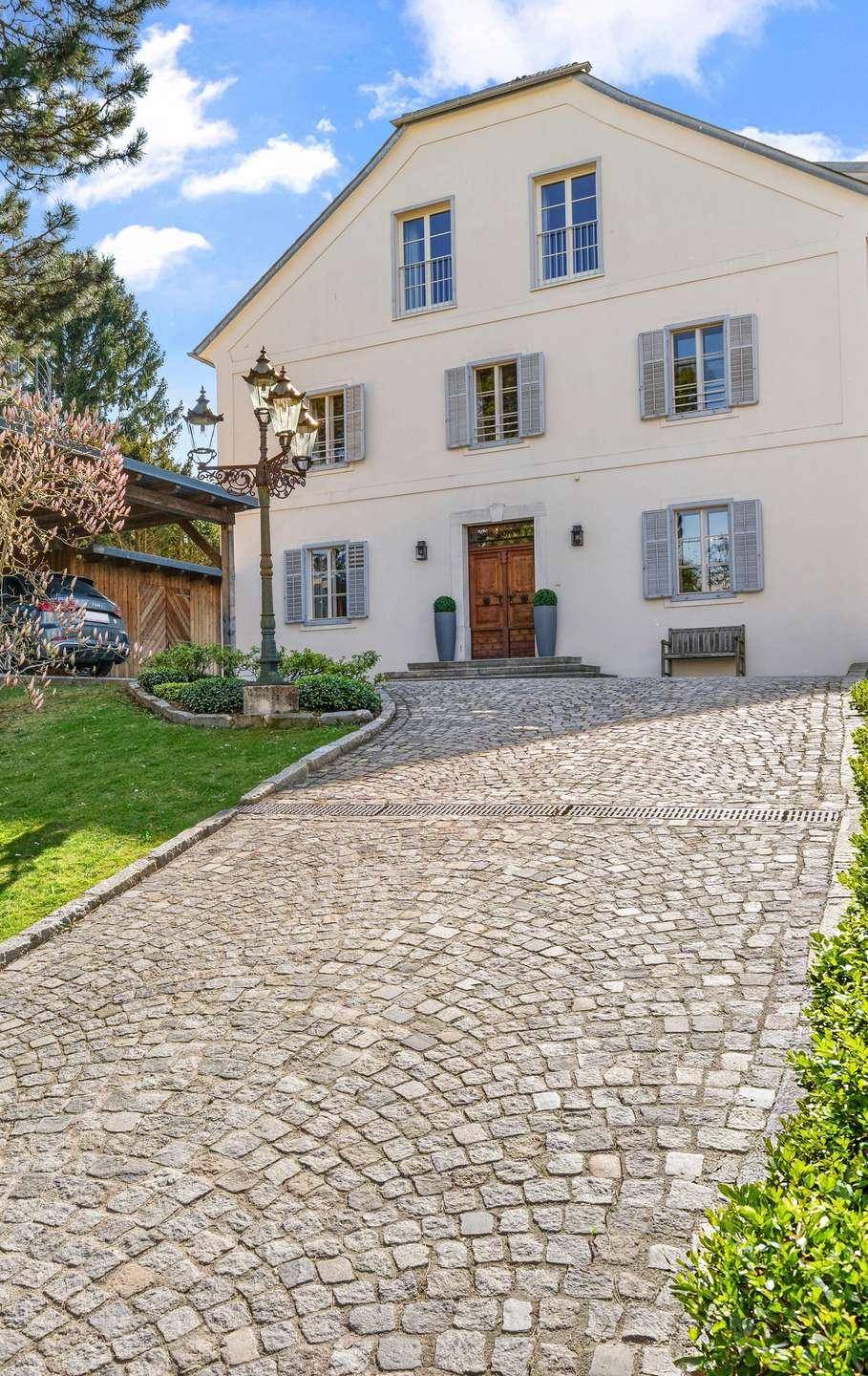 Elegante Hausfassade mit gepflasterter Auffahrt, klassischer Laterne und Carport im Grünen.
