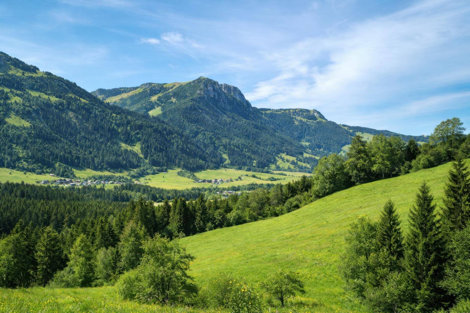 Weite Landschaft mit grünen Hügeln, bewaldeten Bergen und einem Dorf im Tal unter blauem Himmel.