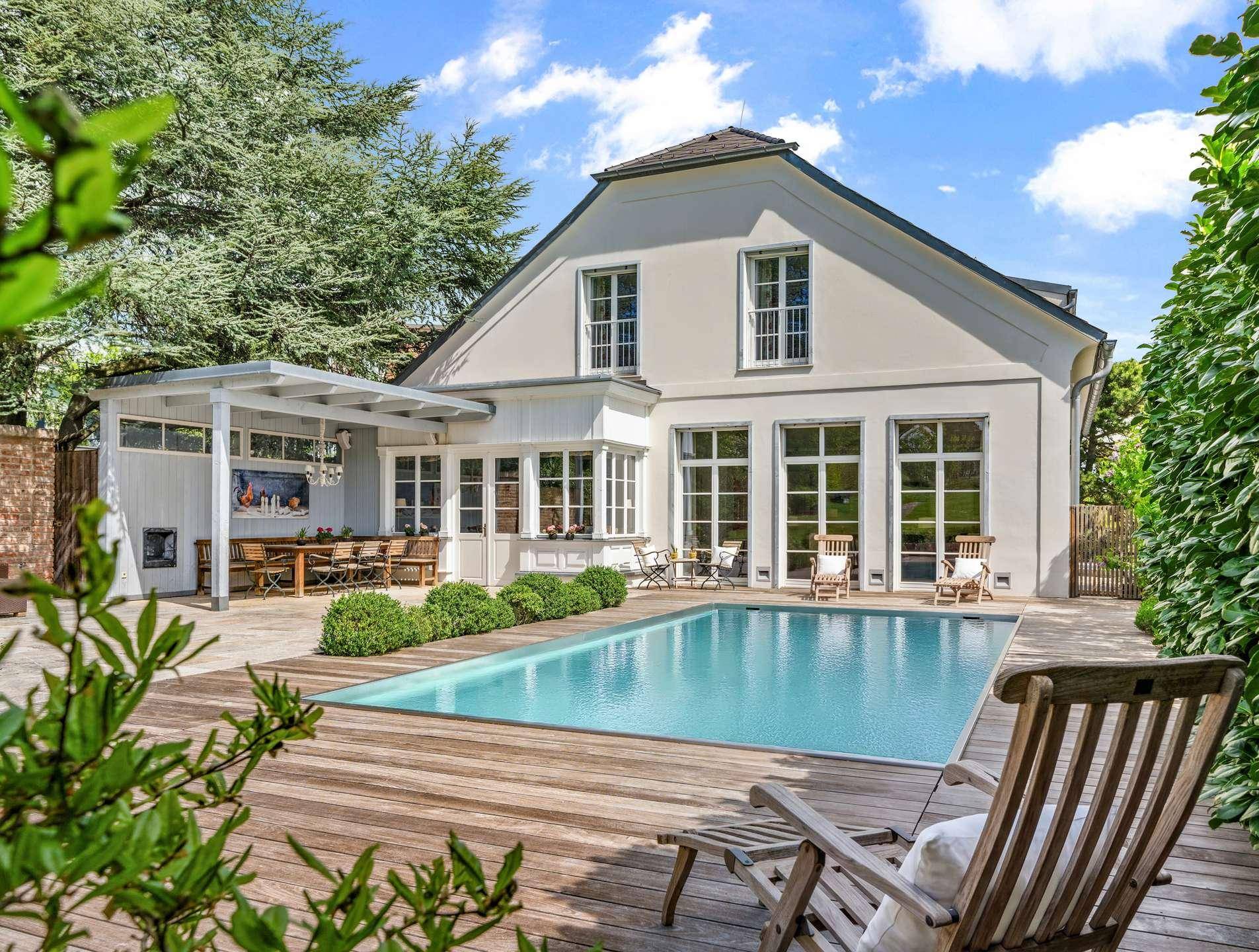 Elegante Villa mit großem Pool, Holzterrasse und gemütlichem Essbereich im Freien, umgeben von Bäumen.