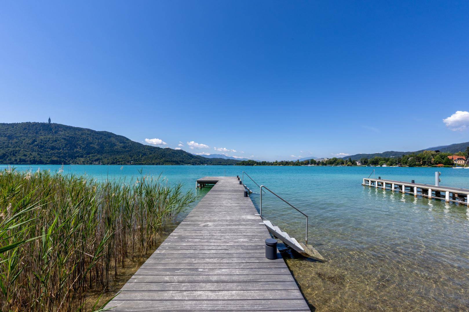 Holzsteg führt ins klare Seewasser, umgeben von Schilf und mit Bergpanorama im Hintergrund.