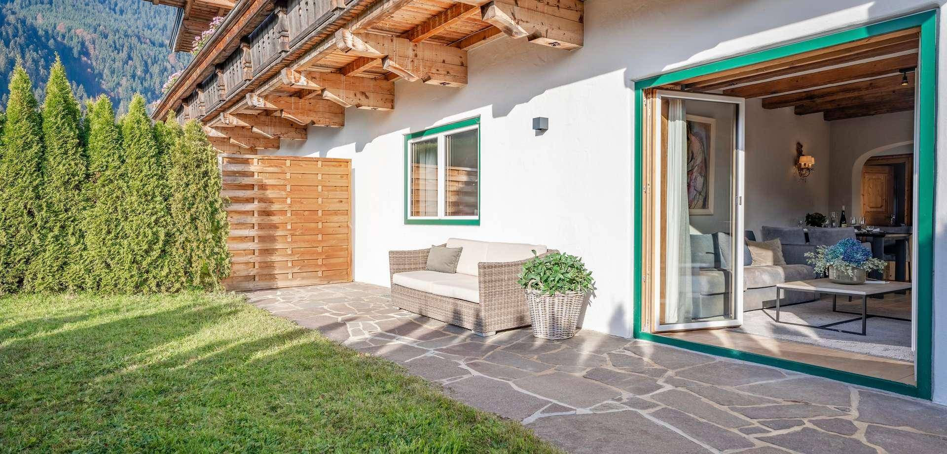 Sonniger Terrassenbereich mit Steinplatten, Gartenmöbeln und Blick auf die Berge.