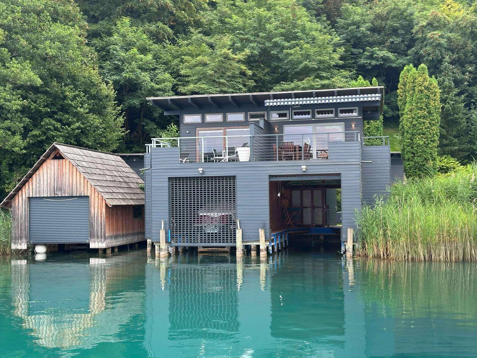 Modernes Haus am Wasser mit Bootsanleger und angrenzendem Holzschuppen, umgeben von Bäumen.