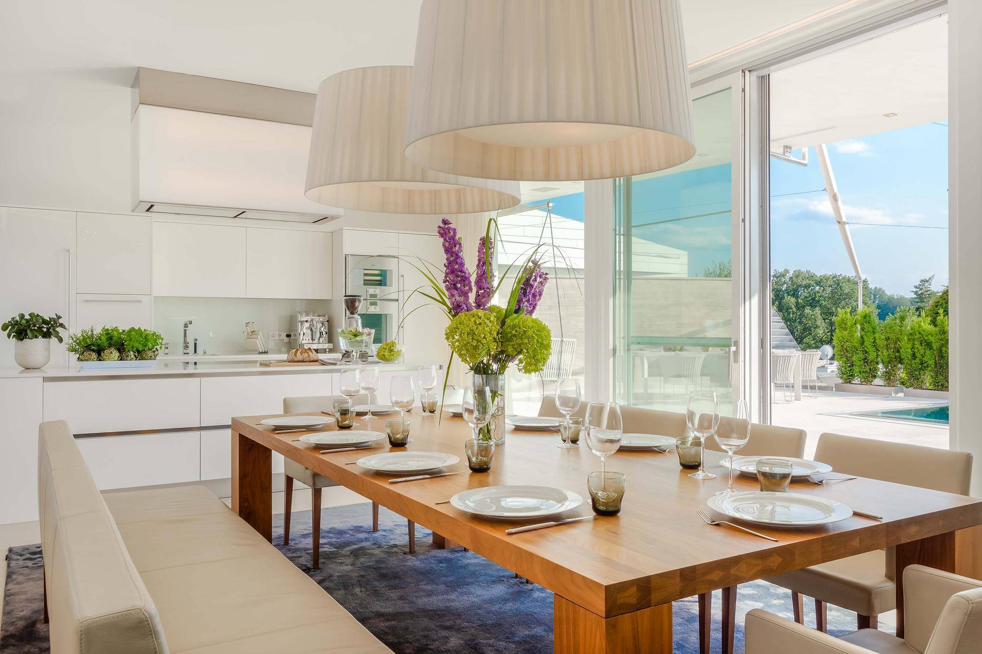 Eleganter Essbereich mit Holztisch und modernen Pendelleuchten, offen zur weißen Küche und mit Zugang zur Terrasse.