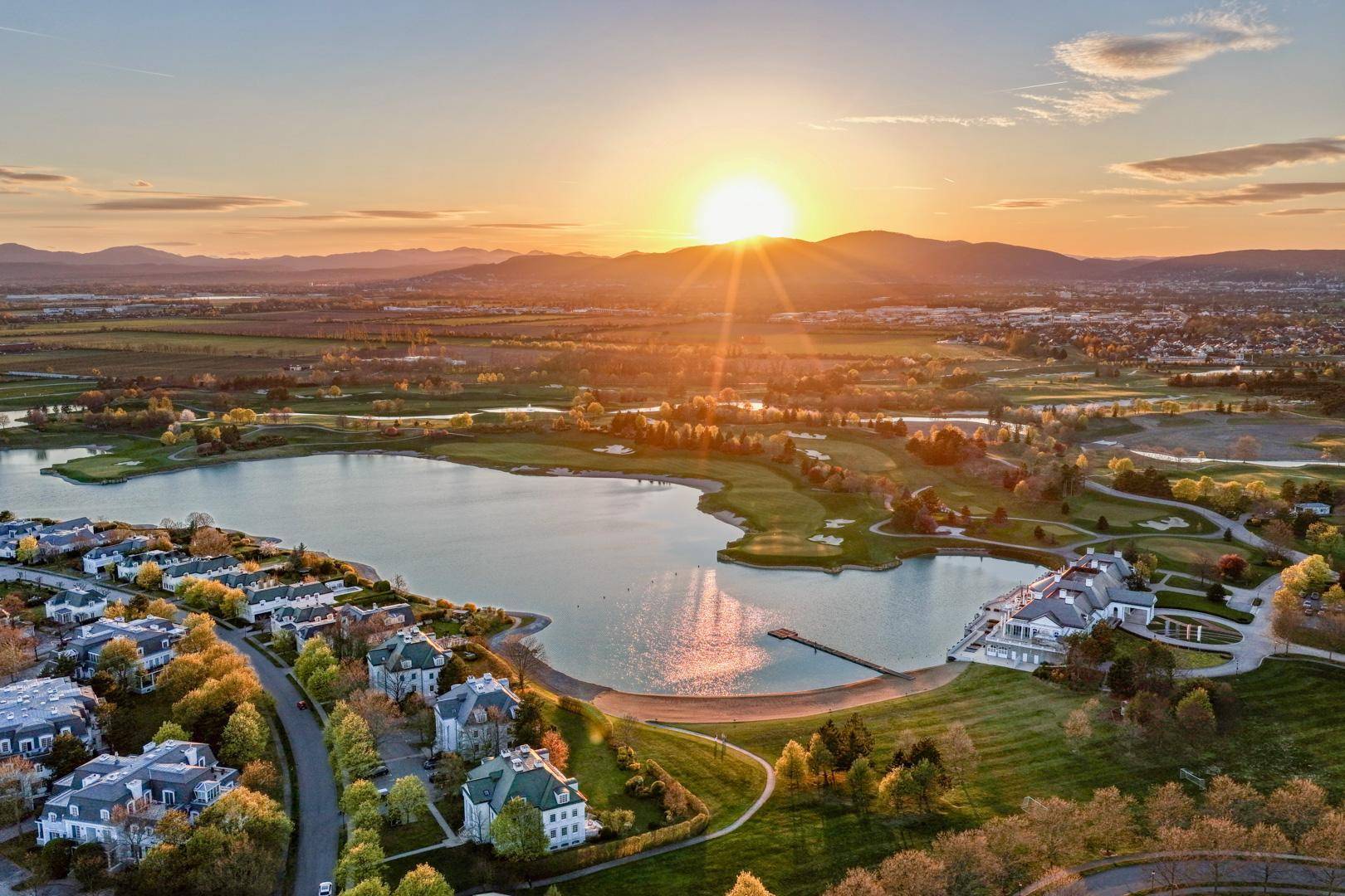 Weitläufige Luftaufnahme der Wohnanlage am Seeufer bei Sonnenuntergang mit Golfplatz und Bergen im Hintergrund.