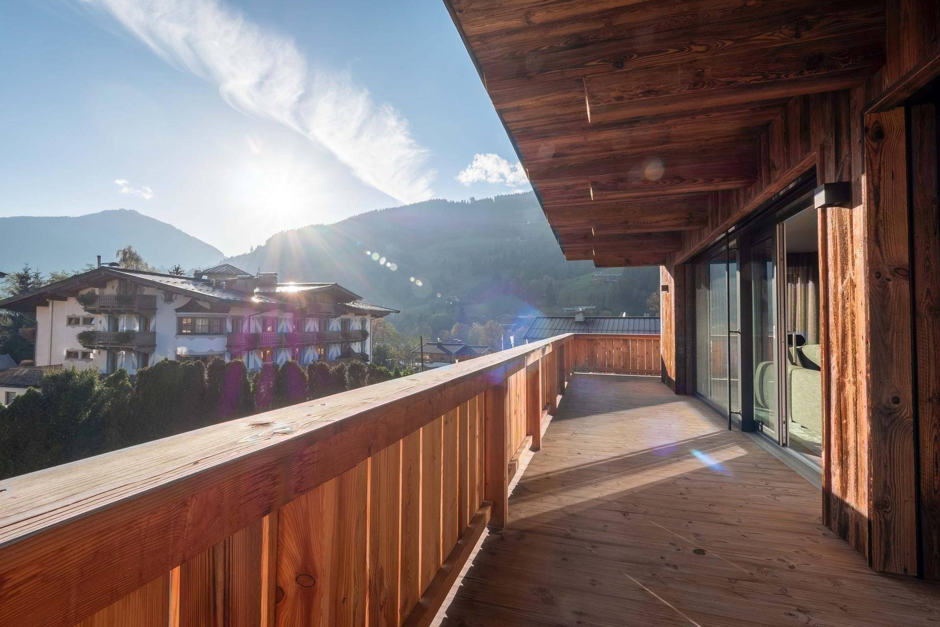Geräumiger Holzbalkon mit sonnigem Ausblick auf die umliegende Berglandschaft und weitere Häuser.