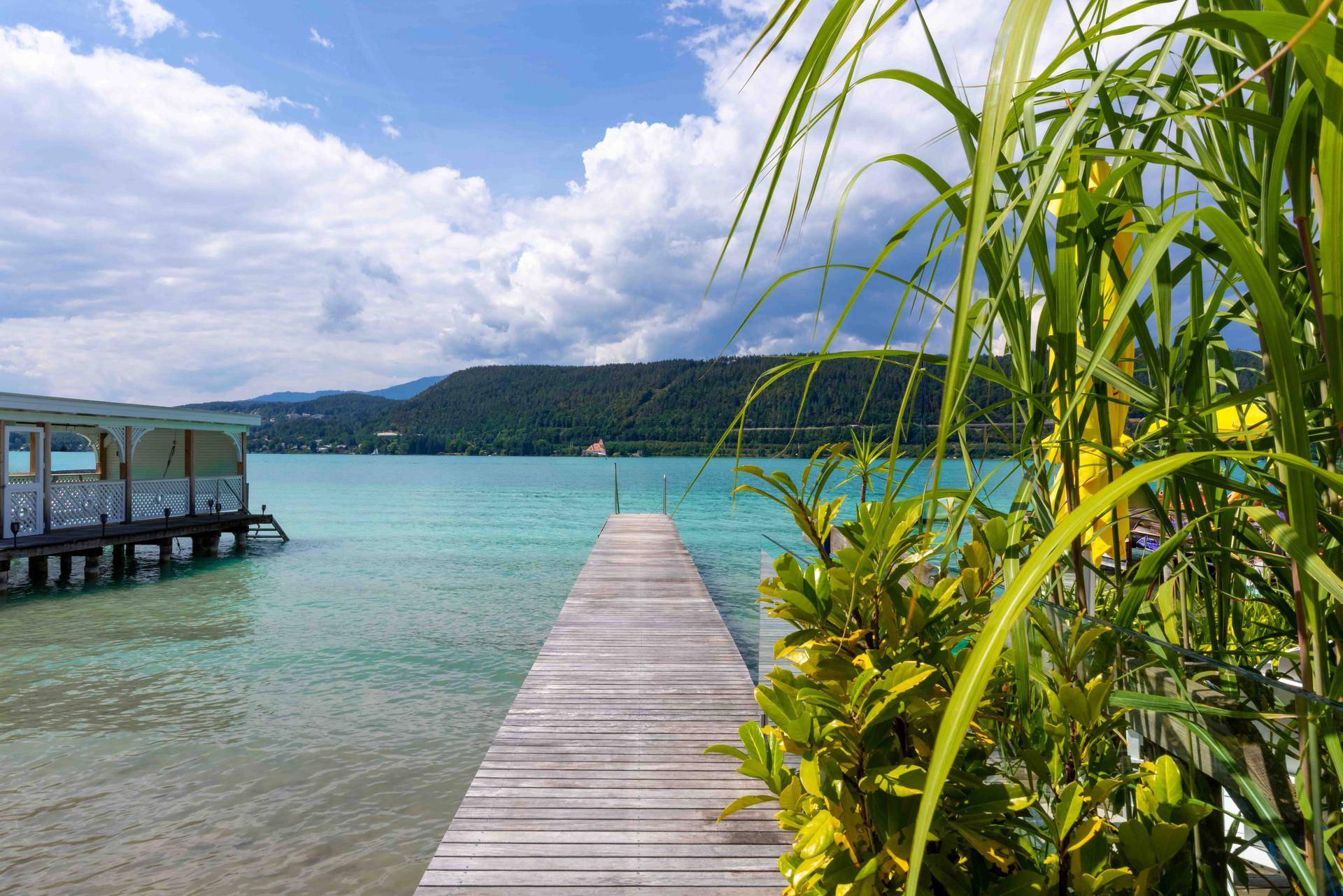 Langer Holzsteg führt in den türkisblauen See, umgeben von grünen Hügeln und einem blauen Himmel.