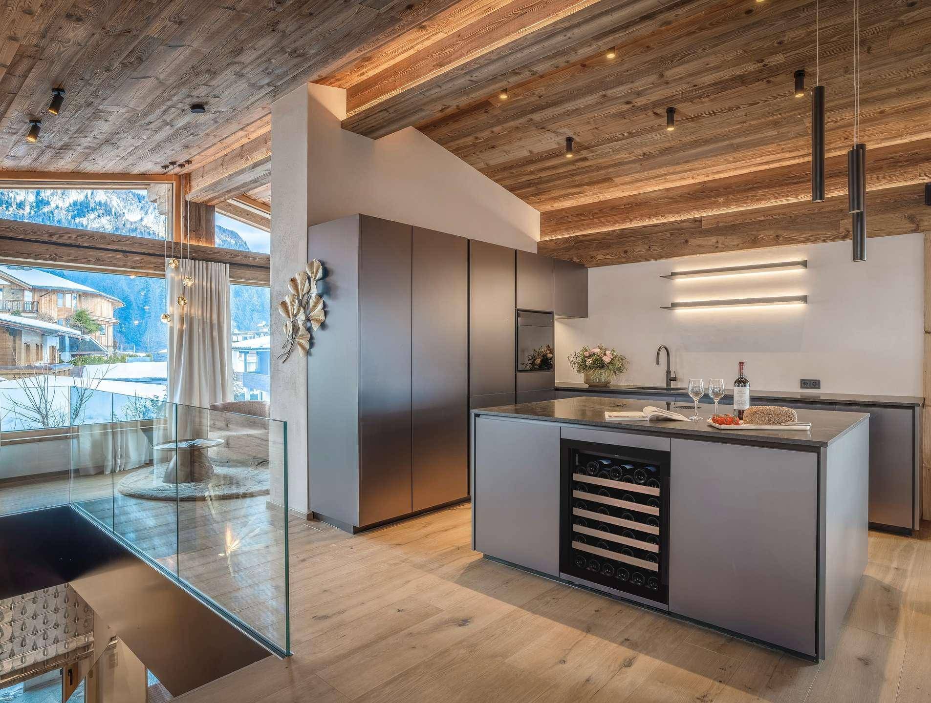 Moderne Küche mit grauen Schränken, Kücheninsel, Weinkühler und viel Tageslicht durch große Fenster.