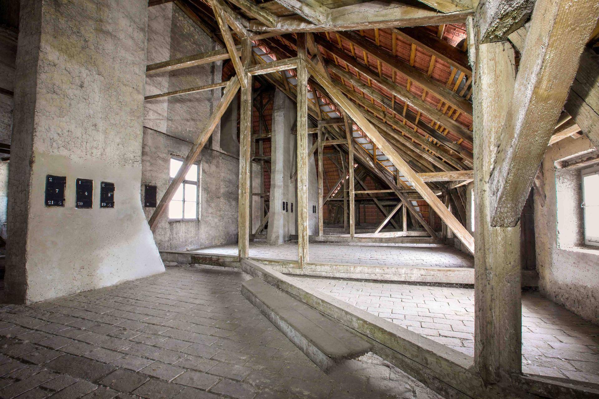 Geräumiger Dachboden mit freiliegenden Holzbalken, Ziegelboden und natürlichem Lichteinfall durch kleine Fenster.