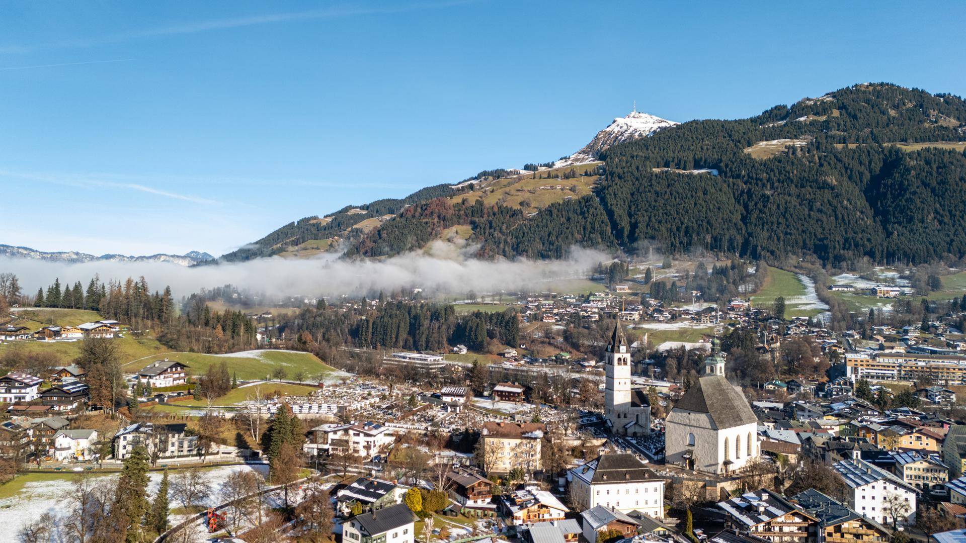 Panoramablick auf eine verschneite Stadt mit Kirche und umliegenden Bergen unter blauem Himmel.