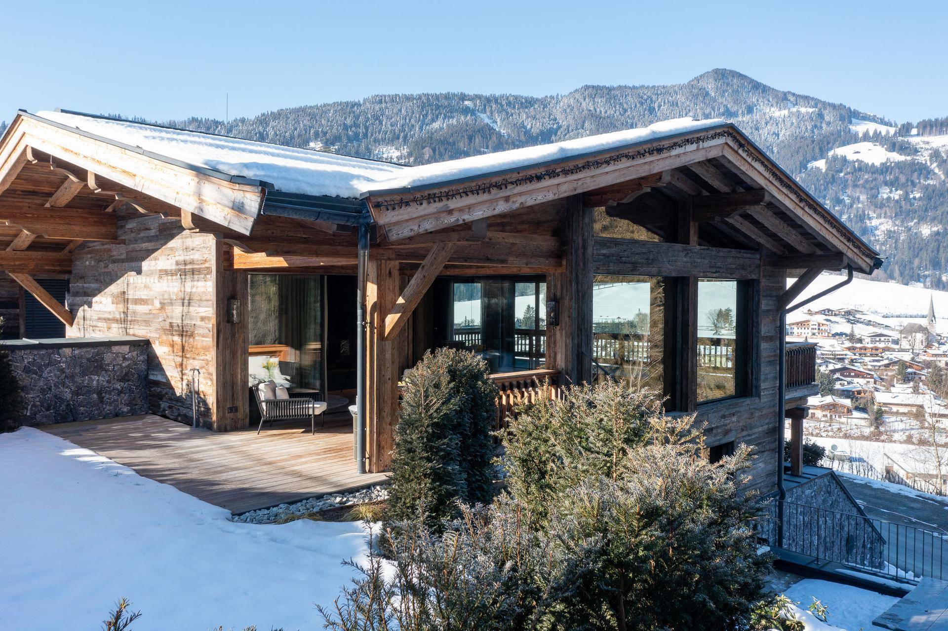 Sonniger Außenbereich mit Holzterrasse, Sitzgelegenheiten und Blick auf die verschneite Landschaft.