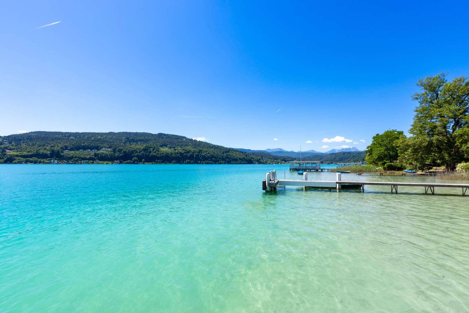 Weitläufiger Blick über den kristallklaren See mit mehreren Anlegestellen und bewaldeten Bergen im Hintergrund.