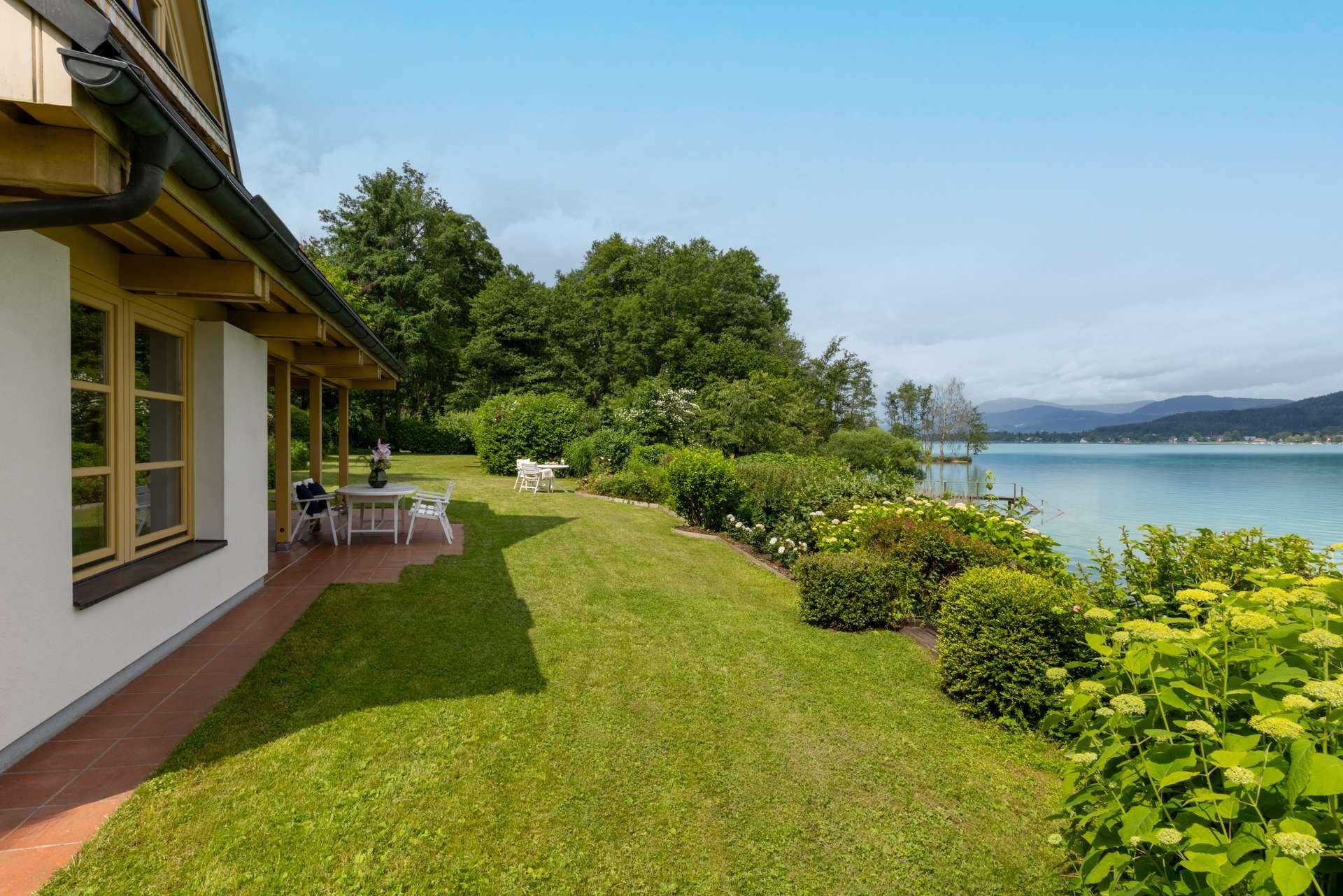 Weitläufiger Garten mit Rasenfläche und Terrasse am Seeufer, umgeben von üppiger Vegetation.