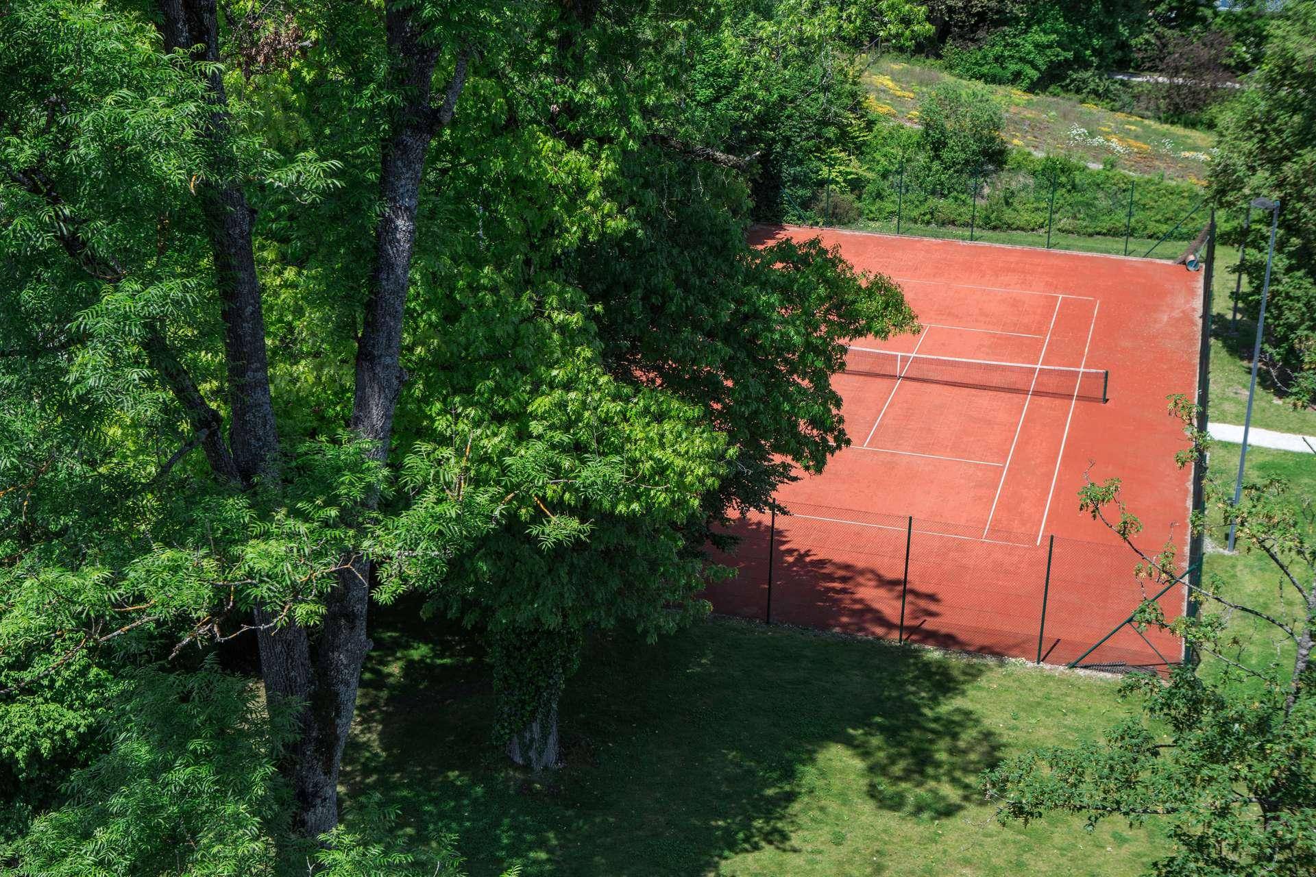 Roter Tennisplatz umgeben von grünen Bäumen und Rasenflächen.