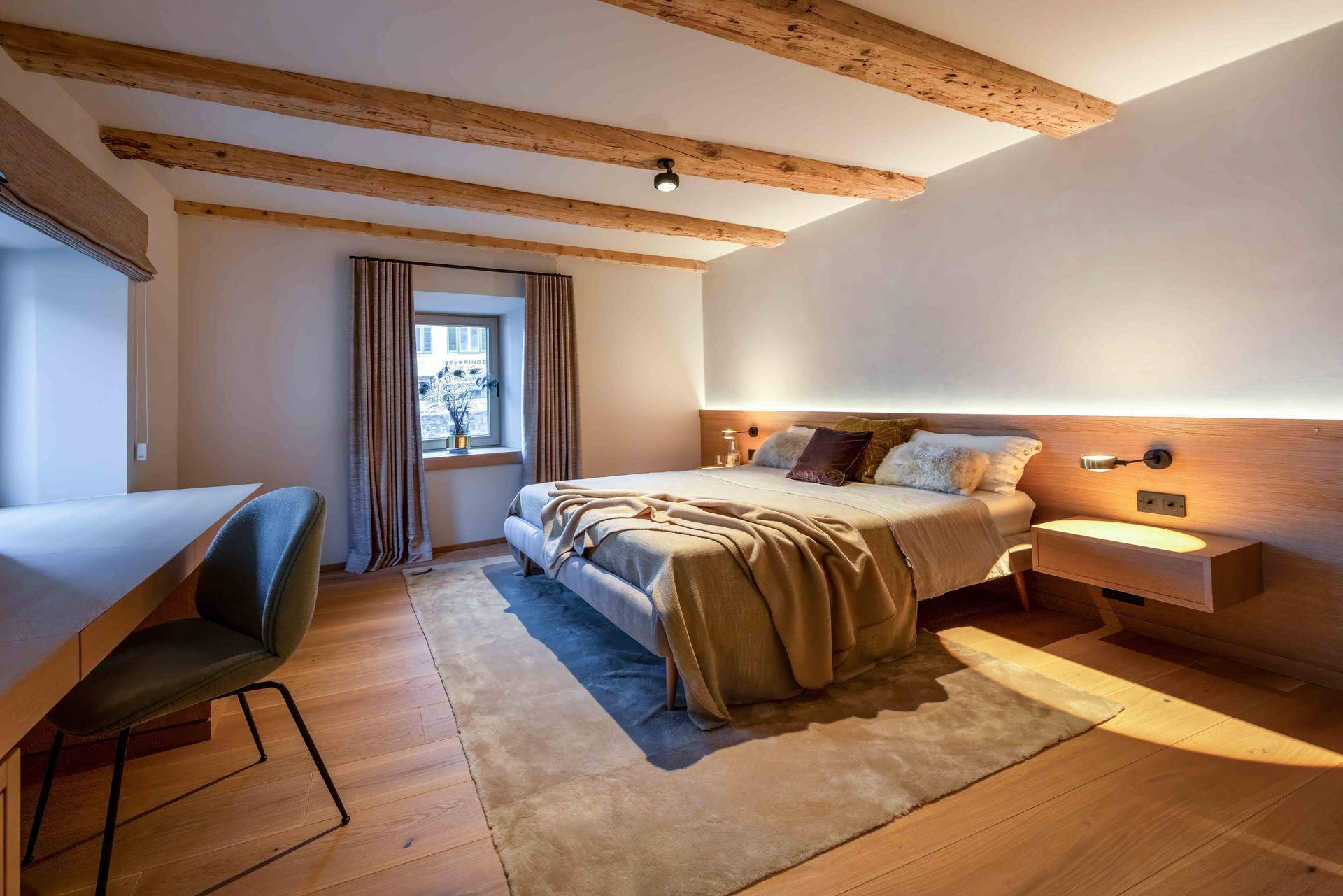 Schlafzimmer mit Holzbalken, großem Bett und Schreibtisch in warmer Atmosphäre.