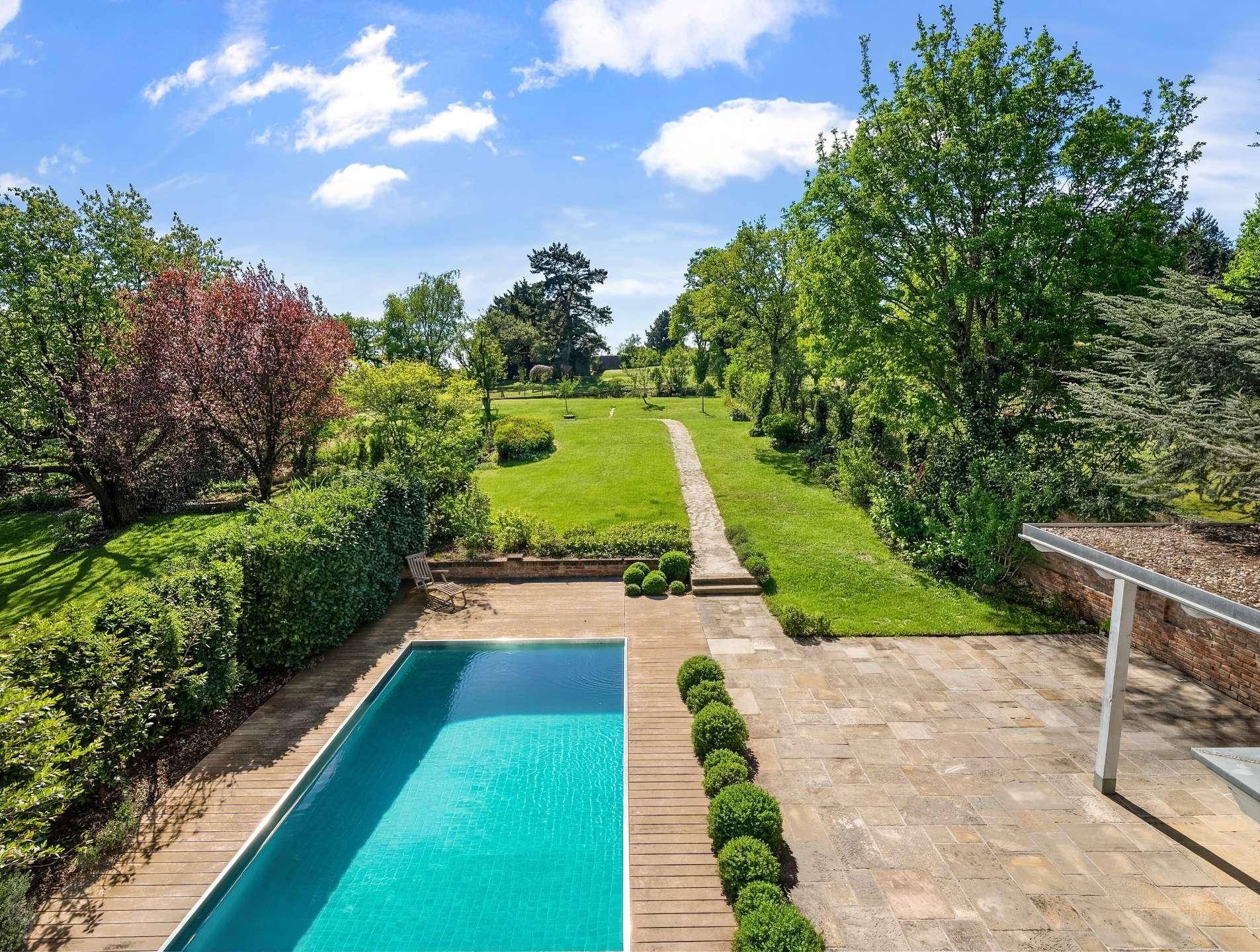 Großer, gepflegter Garten mit modernem Pool, weitläufiger Rasenfläche und altem Baumbestand unter blauem Himmel.