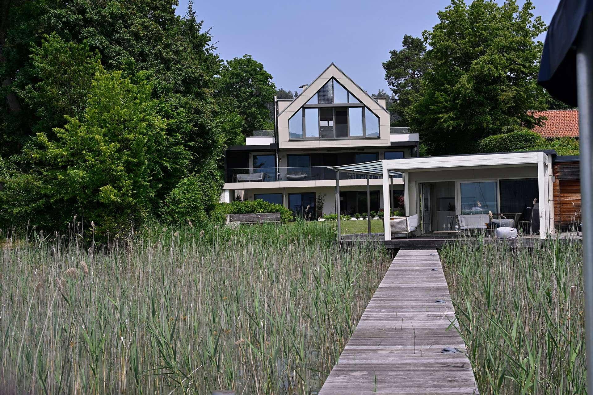Modernes Haus mit Seeblick, umgeben von üppigem Grün und einem Holzsteg im Vordergrund.