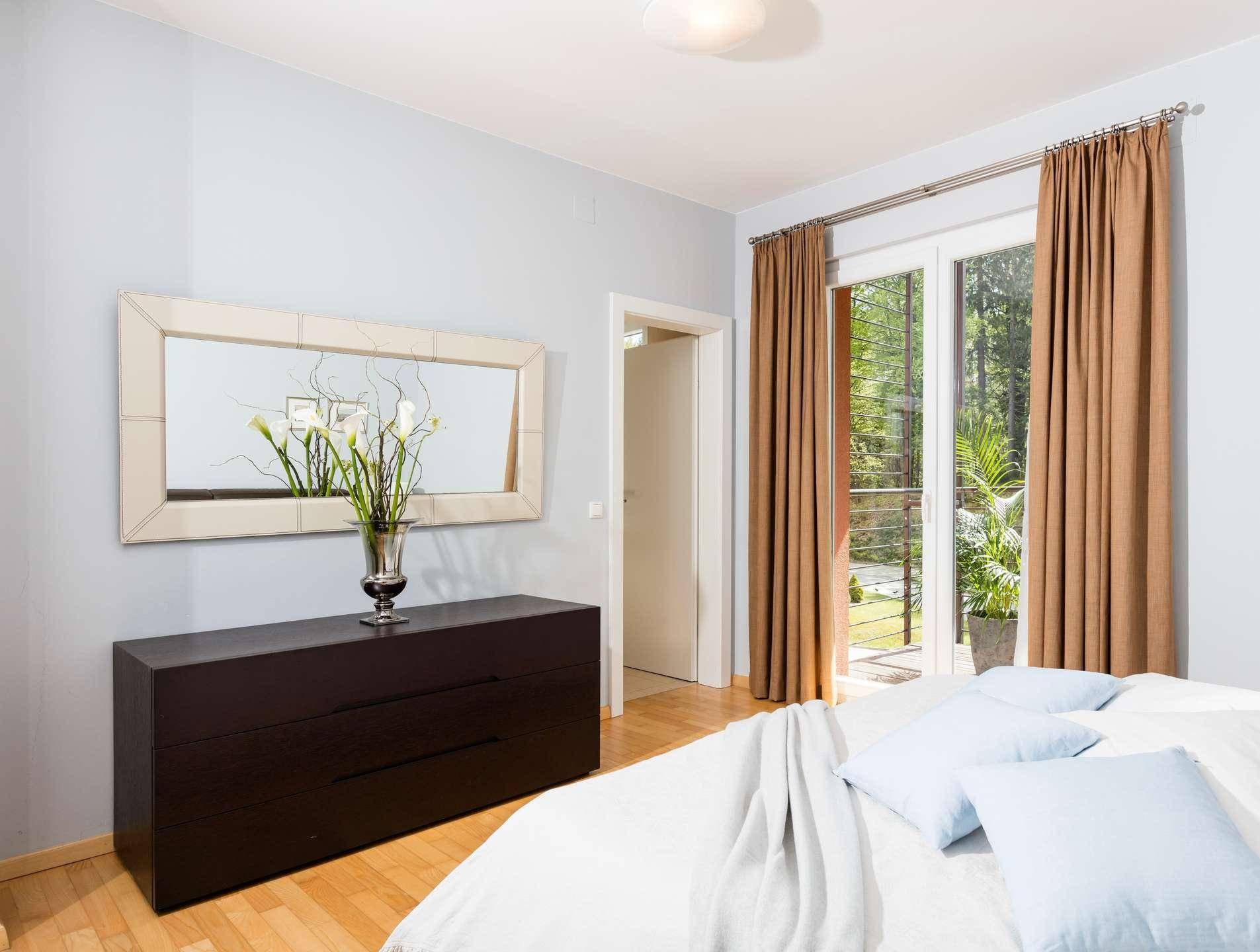 Schlafzimmer mit dunkler Kommode, großem Spiegel und Zugang zu einem grünen Außenbereich durch Glastüren.
