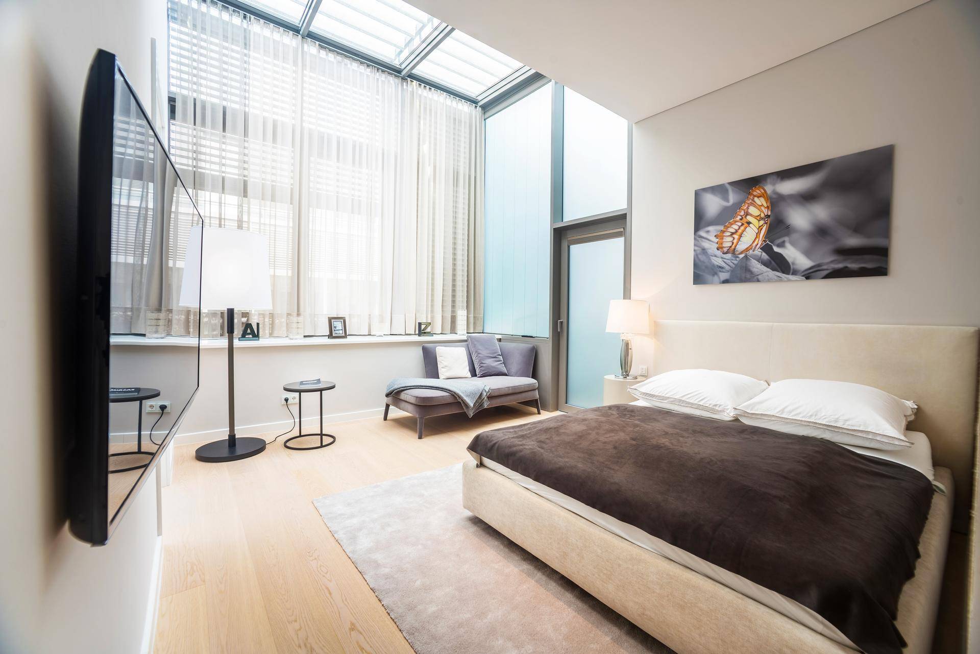 Helles Schlafzimmer mit großem Bett, Wandfernseher und raumhohen Fenstern mit Jalousien.