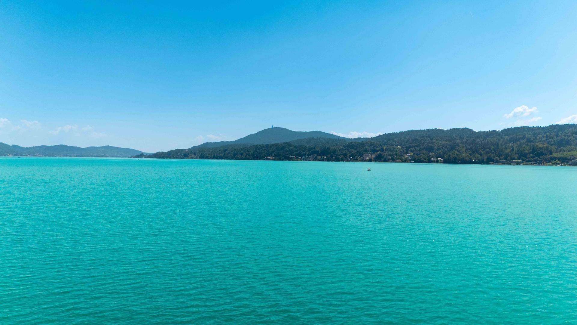 Weitläufiger Blick über den türkisfarbenen See mit bewaldeten Hügeln unter strahlend blauem Himmel.