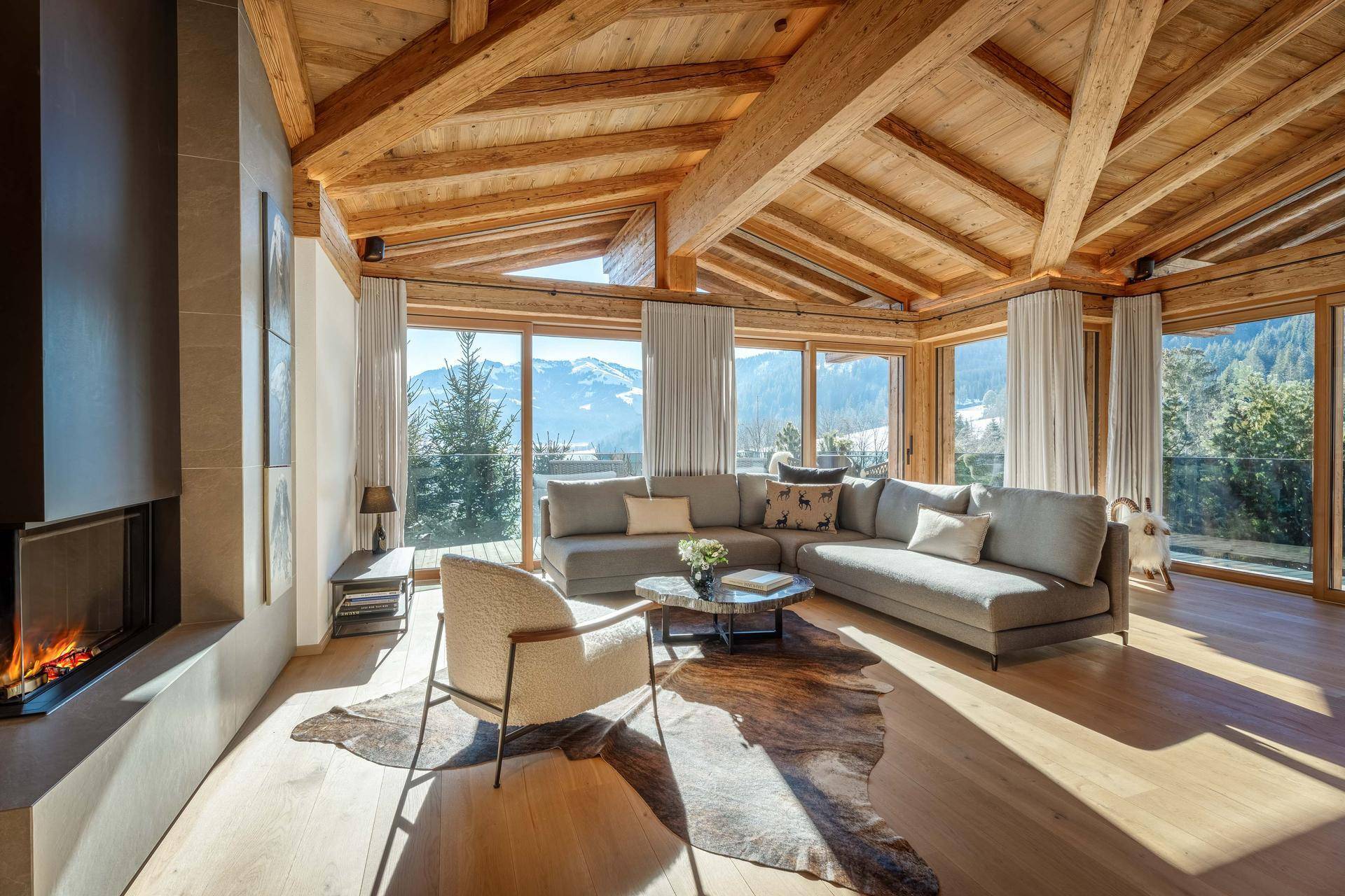 Lichtdurchflutetes Wohnzimmer im alpinen Stil mit Kamin, Holzbalken und Panoramablick auf die Berge.