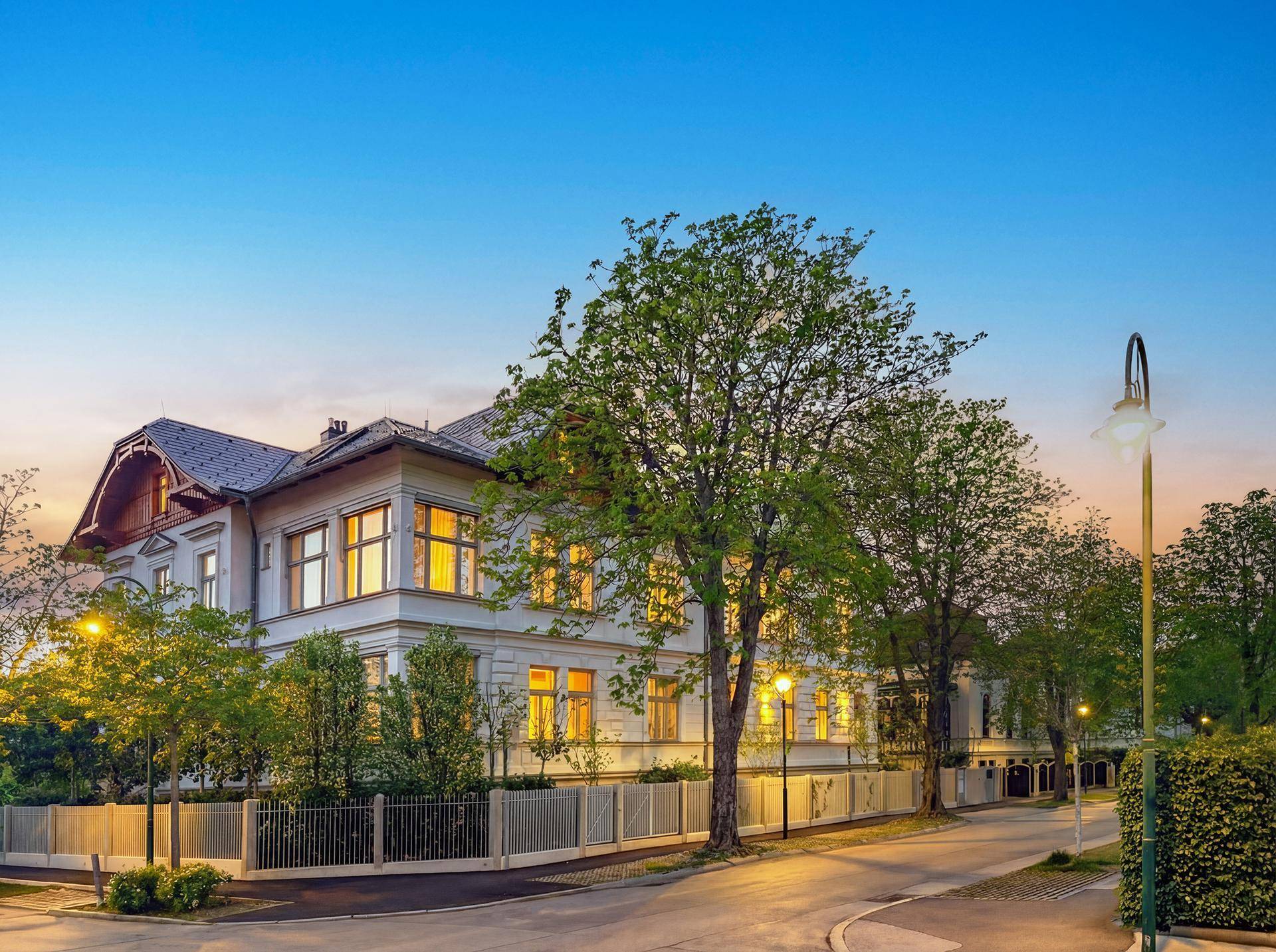 Historische Villa mit beleuchteter Fassade und gepflegtem Vorgarten an einer ruhigen Straße.