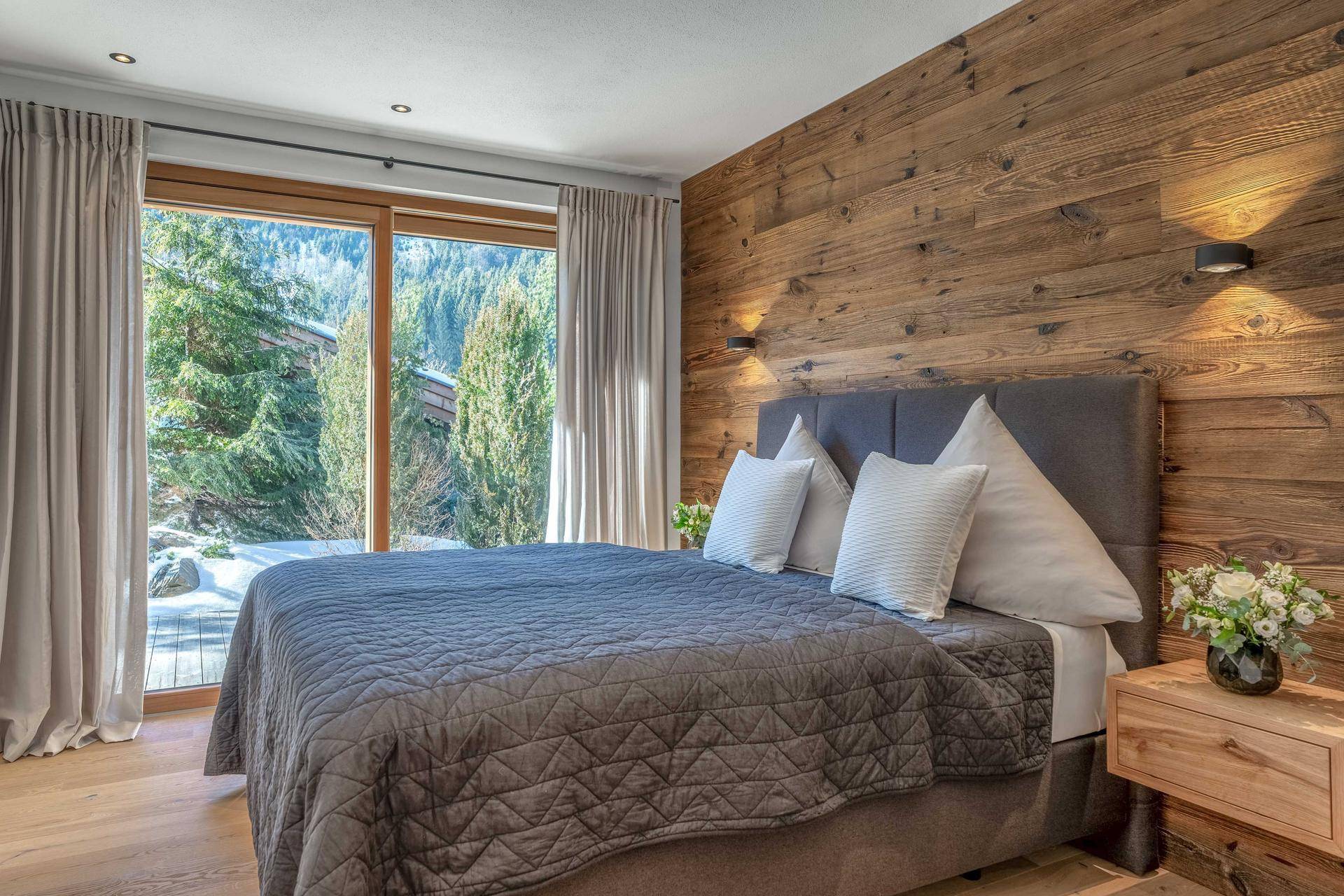 Helles Schlafzimmer mit großen Fenstern, Holzwand und Blick ins Grüne.