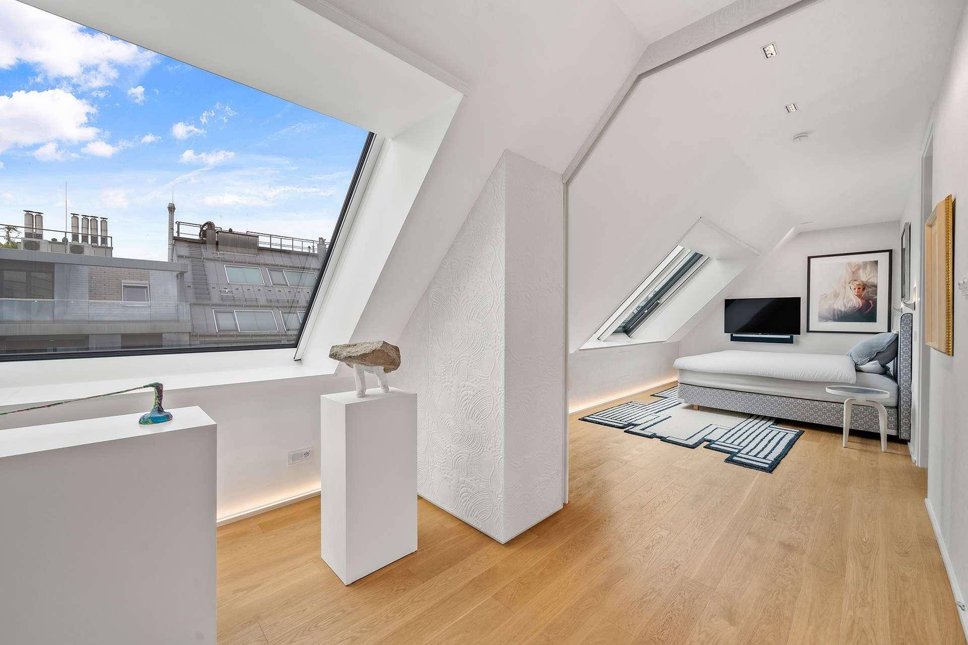 Helles Schlafzimmer im Dachgeschoss mit großen Fenstern, Holzboden und Blick auf die Stadt.