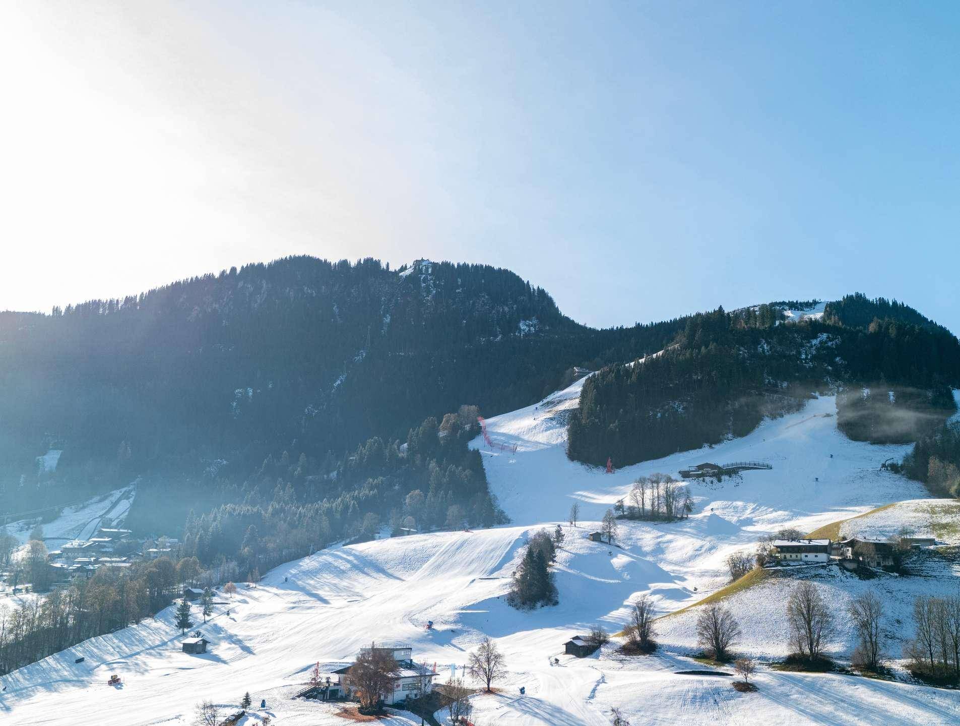 Weitläufige, schneebedeckte Skipiste in den Alpen mit bewaldeten Hängen unter blauem Himmel.