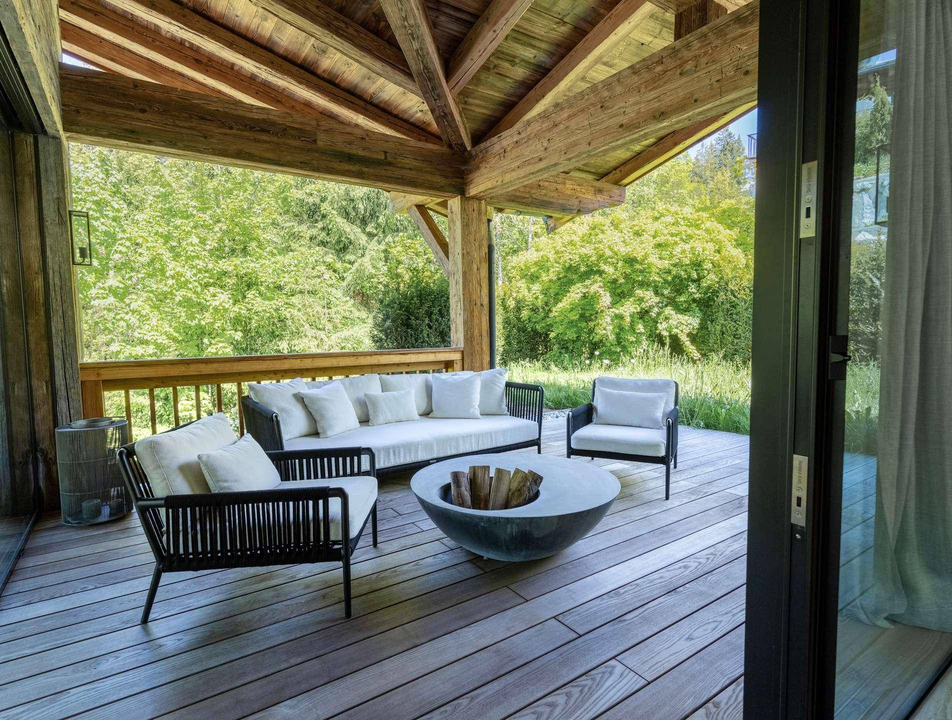 Überdachte Terrasse mit bequemen Loungemöbeln und Feuerstelle, umgeben von üppigem Grün.