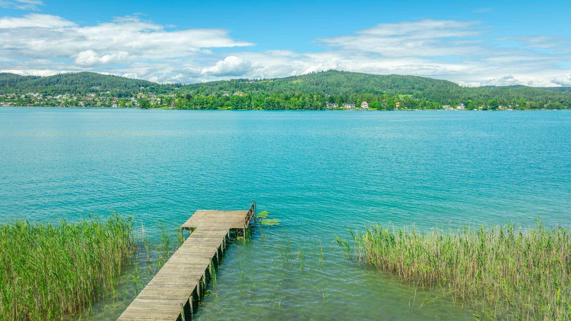 Blick auf einen Holzsteg, der in einen klaren, türkisfarbenen See mit Schilf und bewaldeten Hügeln führt.
