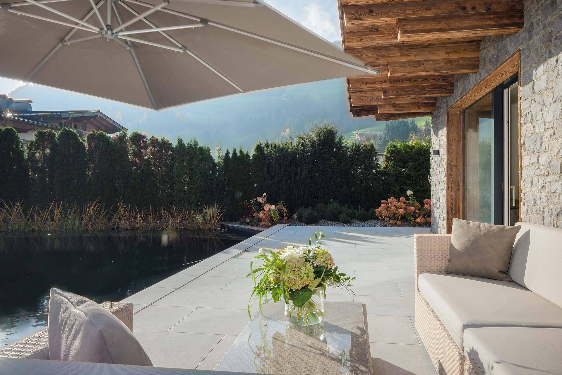 Gemütliche Terrasse mit bequemen Loungemöbeln, großem Sonnenschirm und Blick auf den Naturpool und die Berge.