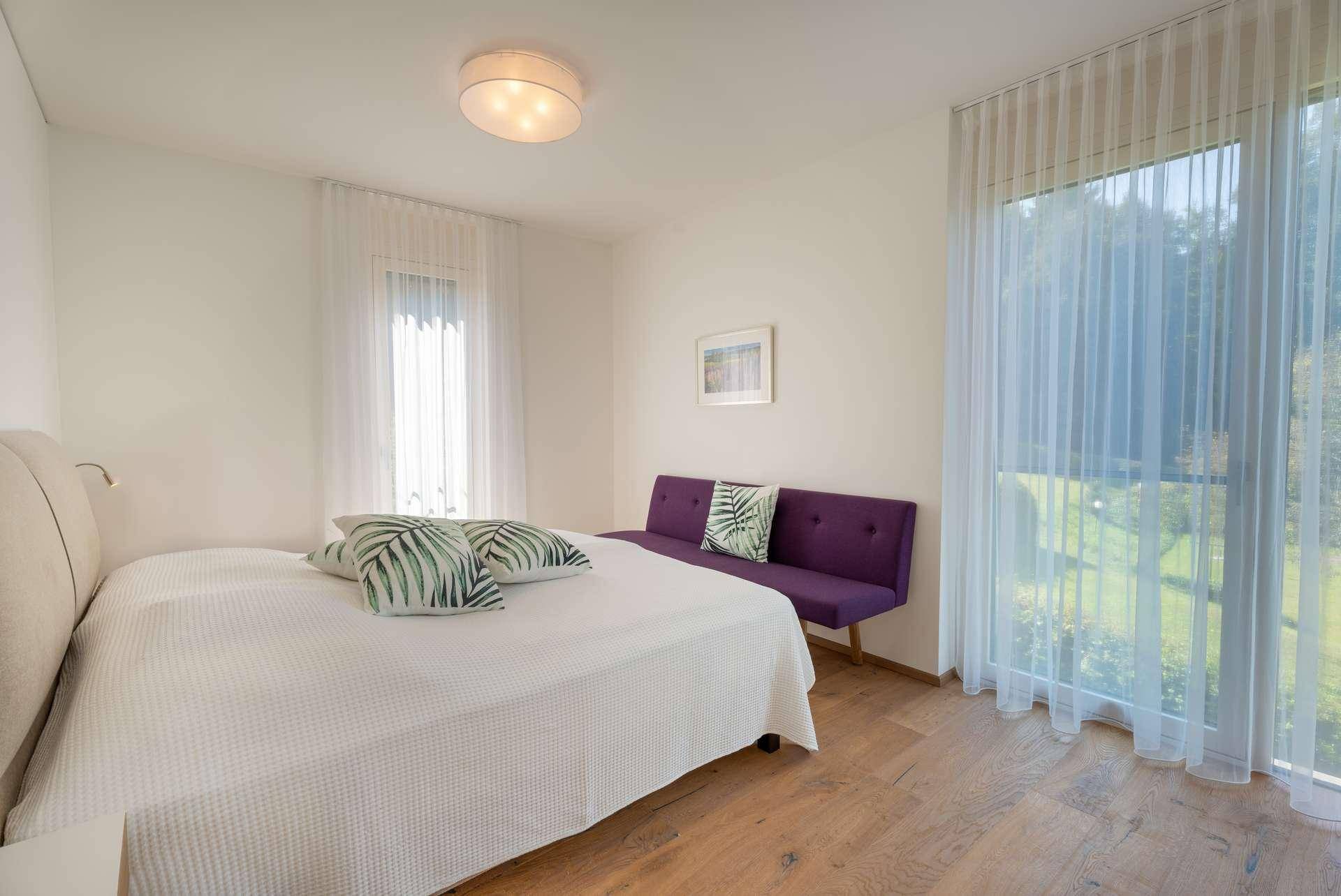 Helles Schlafzimmer mit Holzboden, großem Bett und bodentiefen Fenstern mit Blick ins Grüne.