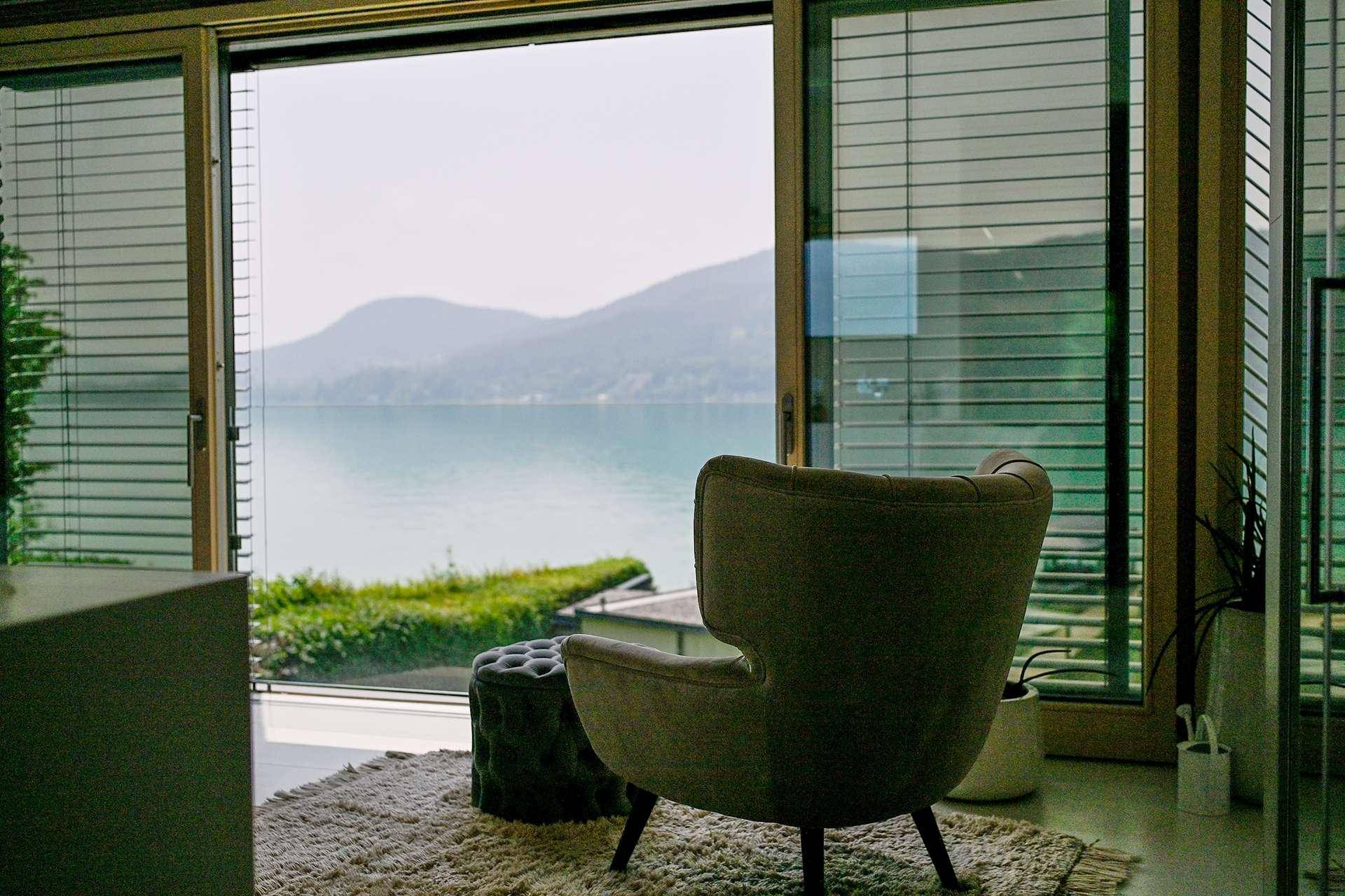 Gemütlicher Sessel am Fenster mit Jalousien und einem weiten Blick über den See und die Berge.