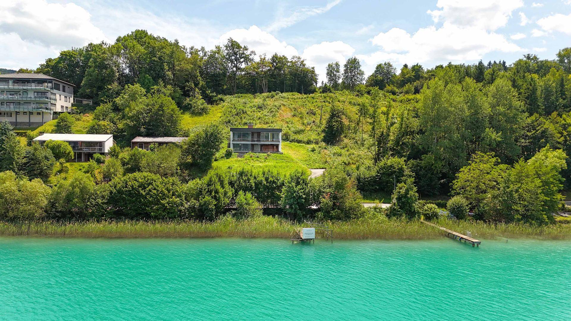 Luftaufnahme eines modernen Hauses am Hang mit direktem Zugang zum türkisfarbenen See und umgebender Natur.