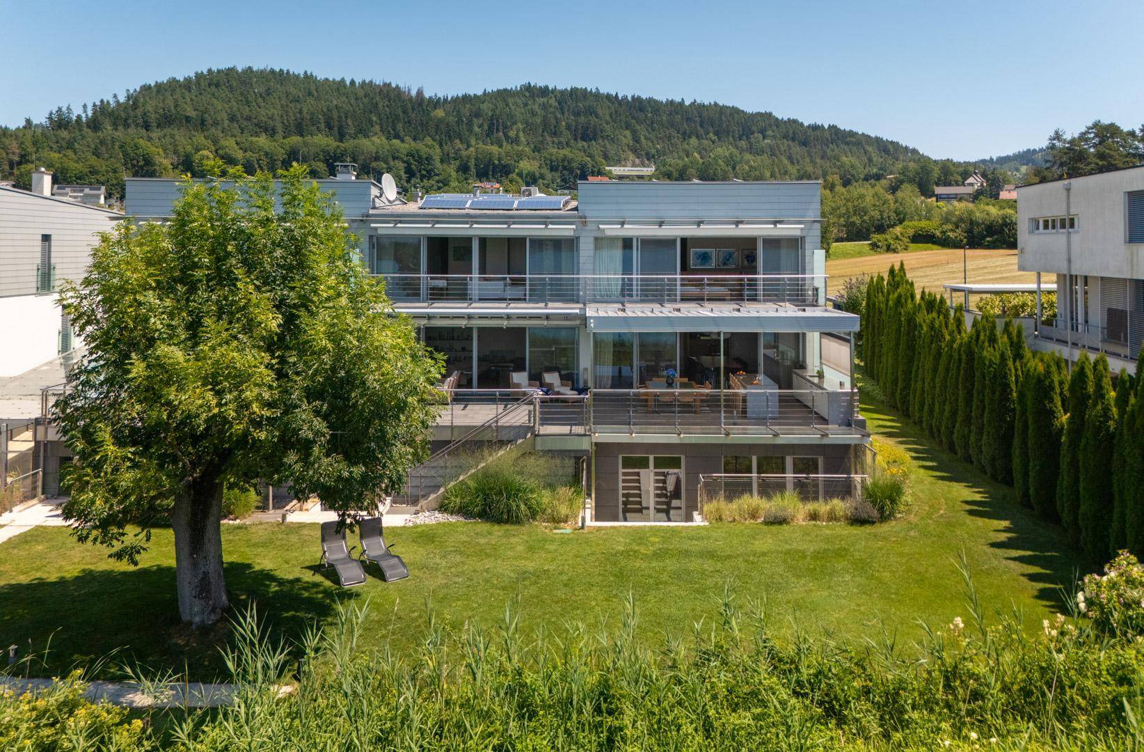Moderne Wohnanlage mit großen Glasfronten, Balkonen und gepflegtem Gartenbereich vor bewaldetem Hügel.