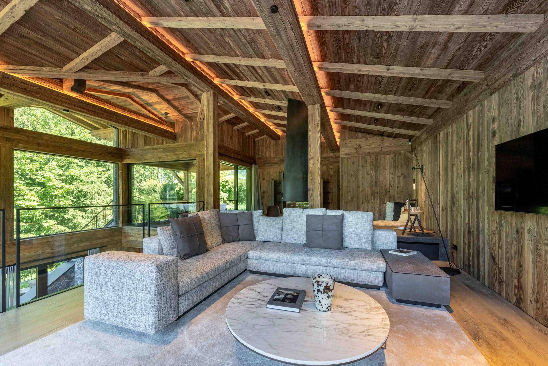 Gemütliches Wohnzimmer im Chalet-Stil mit Holzbalkendecke, modernem Sofa und Blick ins Grüne.
