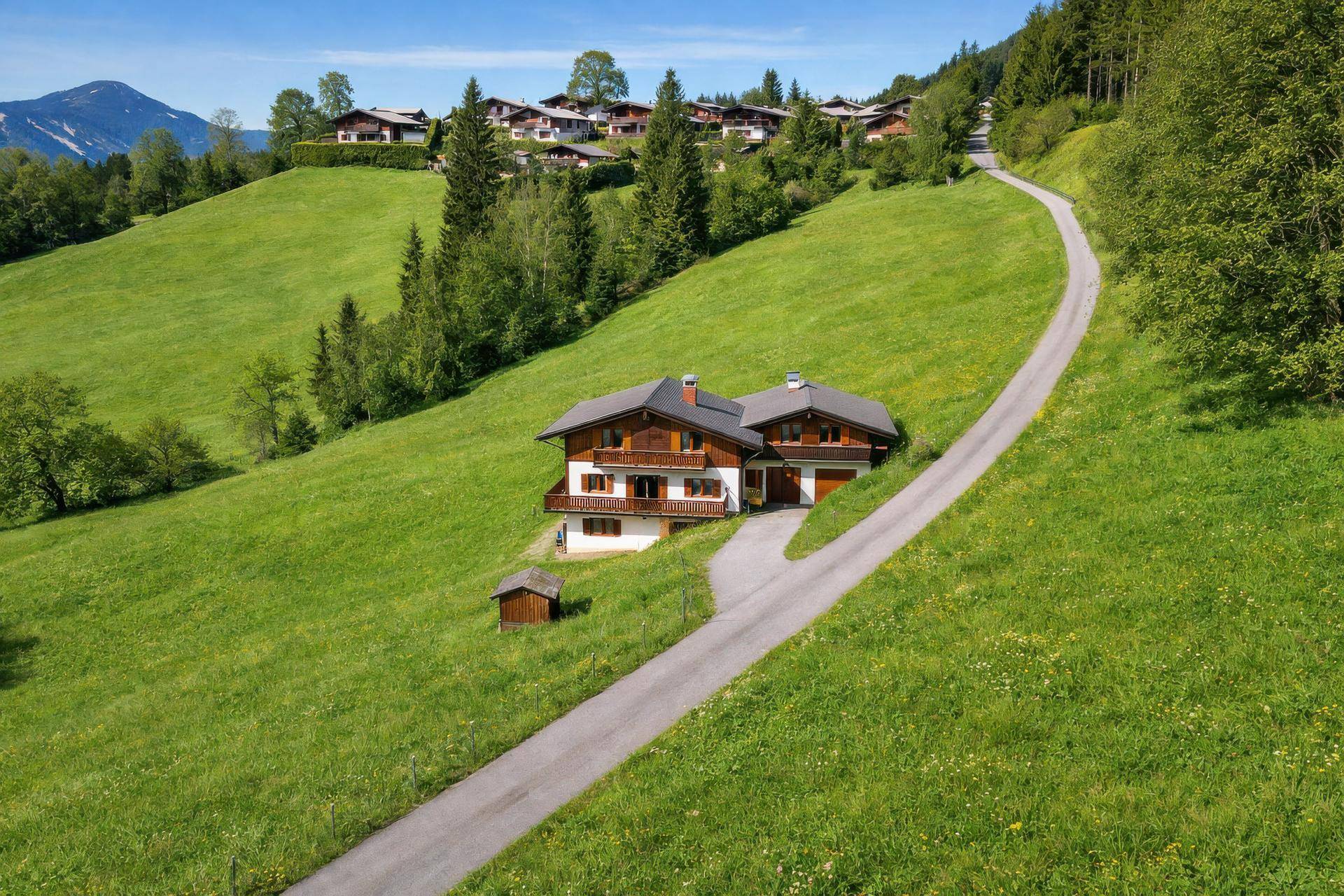 Alpines Haus auf einem grünen Hang mit asphaltierter Zufahrt und weitem Blick auf die Landschaft.