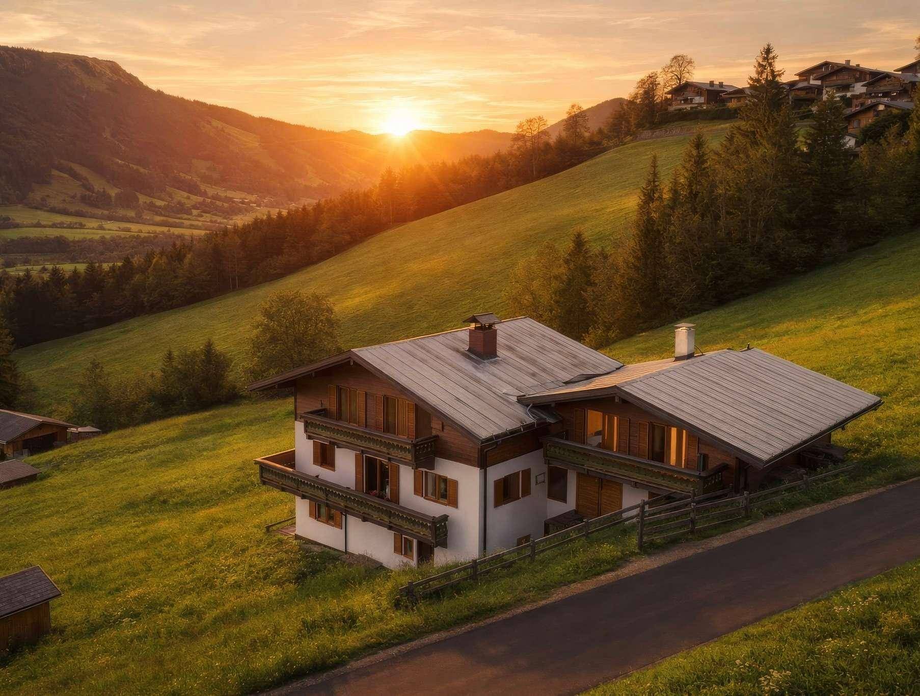 Haus im Alpenstil auf einem grünen Hang bei Sonnenuntergang mit Balkonen und Bergblick.