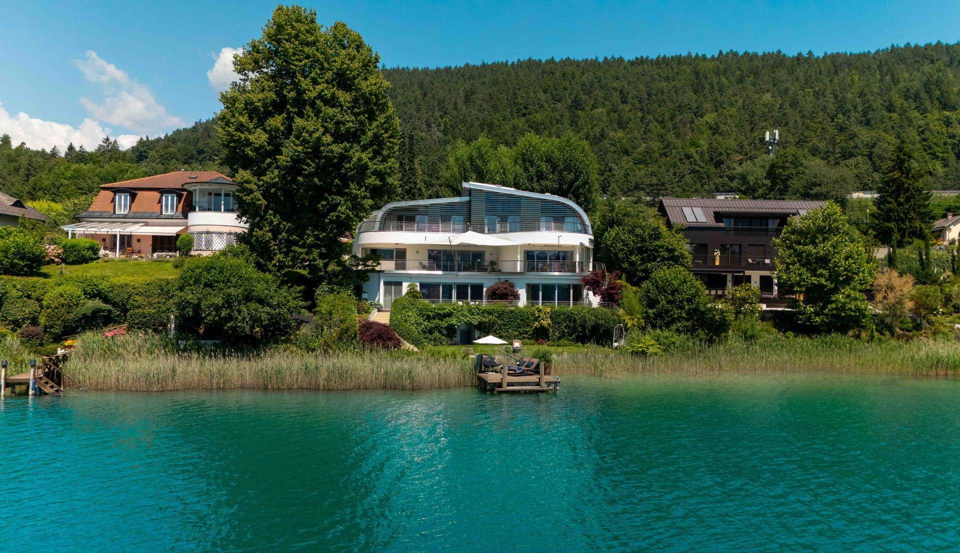 Luftaufnahme eines modernen Hauses direkt am Seeufer, umgeben von Natur und weiteren Gebäuden.