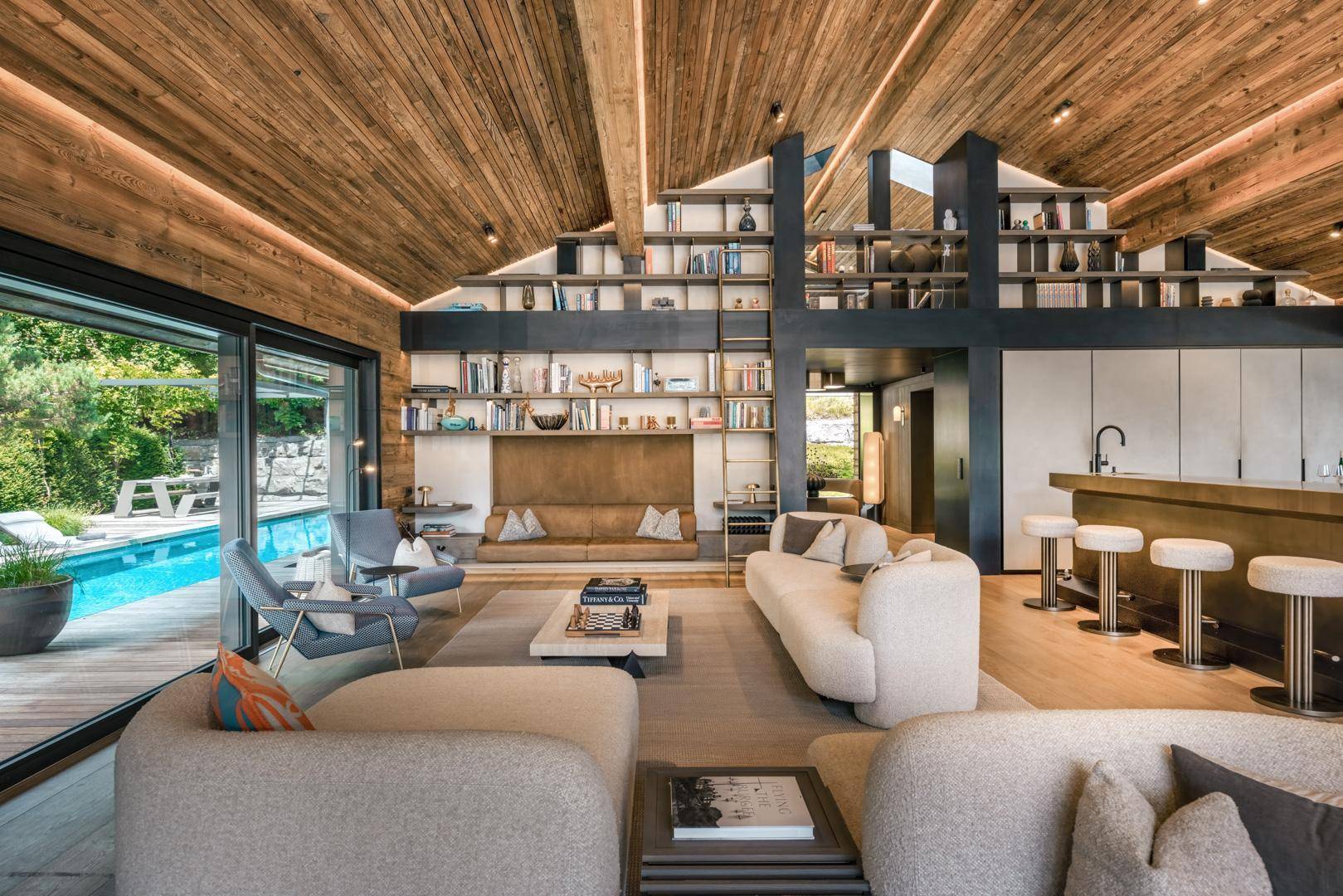 Geräumiges Wohnzimmer mit hoher Holzdecke, integrierten Bücherregalen und Blick auf den Außenpool.
